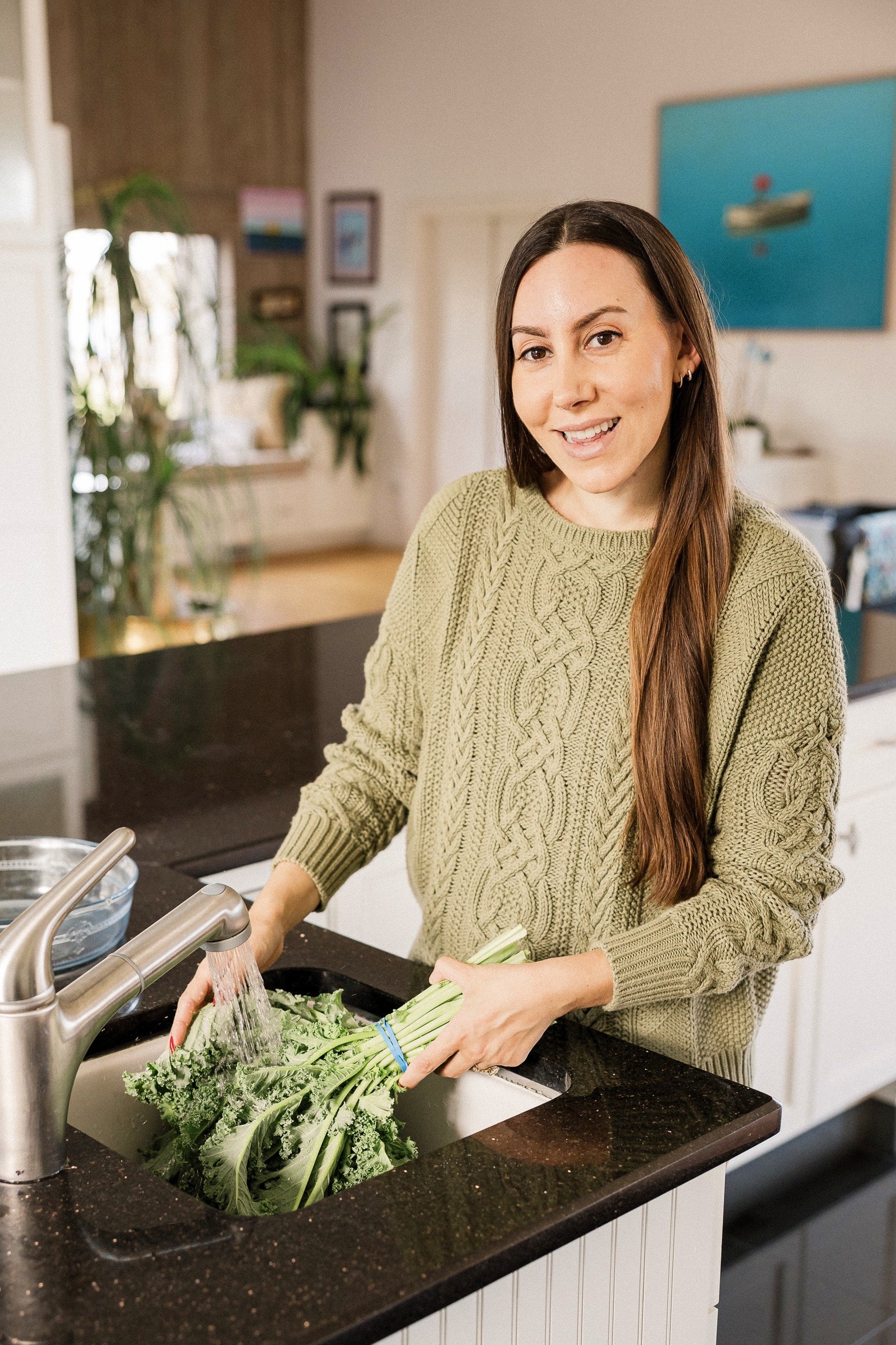 A woman in a green sweater washing kale at the kitchen sink with a mining sink with water running.