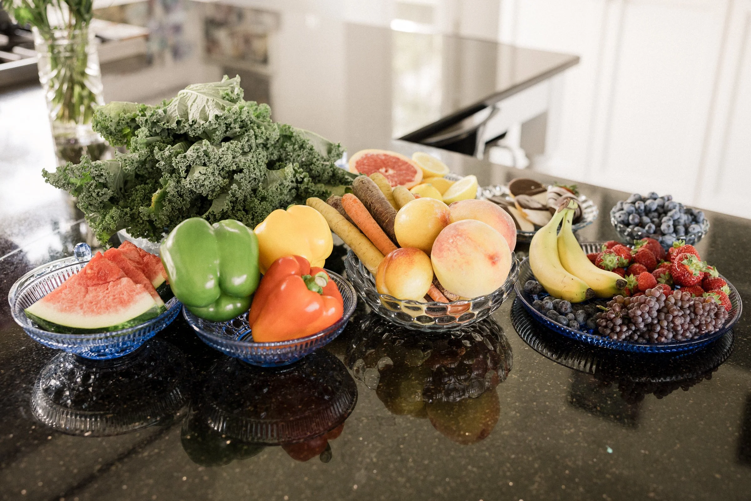 A display of fresh fruits and vegetables on a black granite countertop with glass bowls and plates, including leafy kale, apples, bananas, strawberries, grapes, peaches, watermelon, orange slices, and colorful bell peppers.