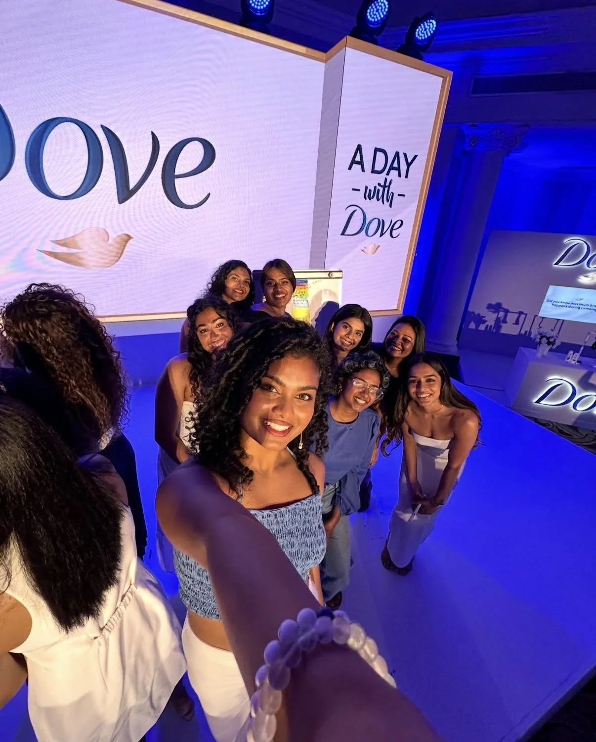 A group of women smiling and taking a selfie at an event with a large screen in the background that reads 'A DAY with Dove'.