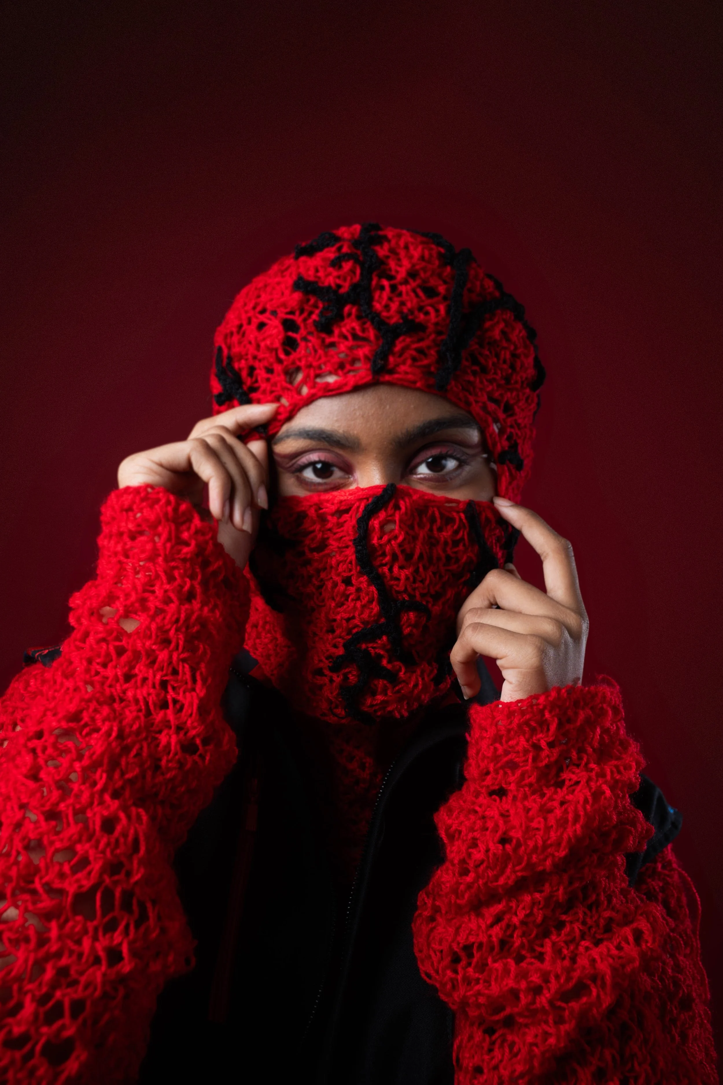 Person with dark skin wearing red crocheted hat, face mask, and matching gloves, covering mouth with fingers, against a dark red background.
