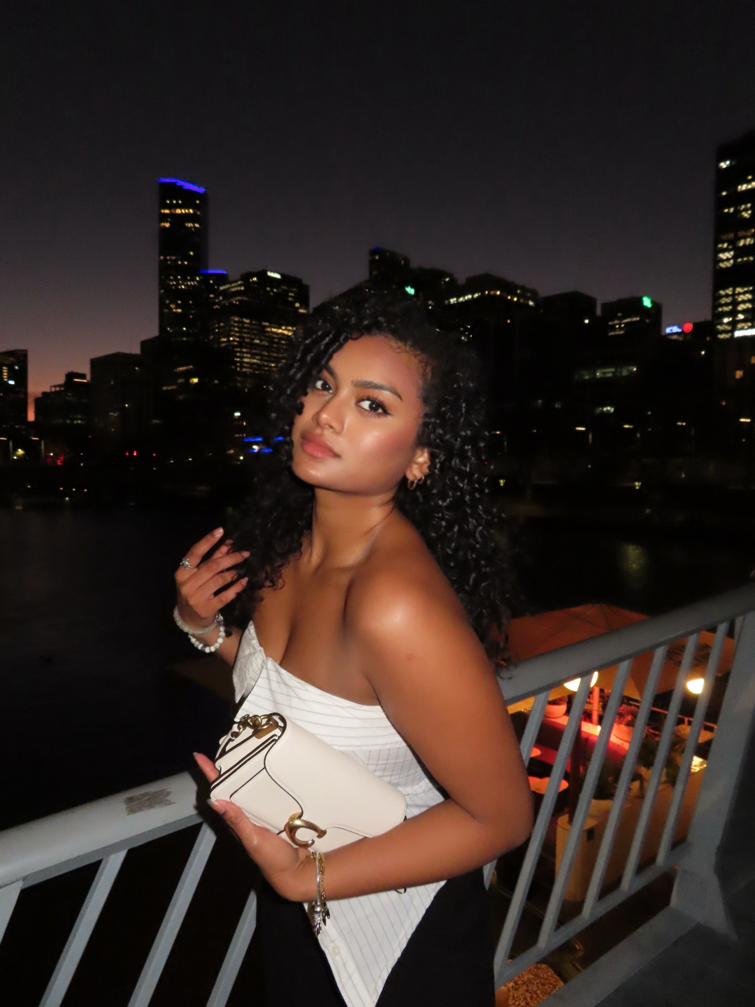 A young woman with curly black hair posing on a balcony at night with city skyscrapers in the background, holding a white handbag and wearing a strapless white top.