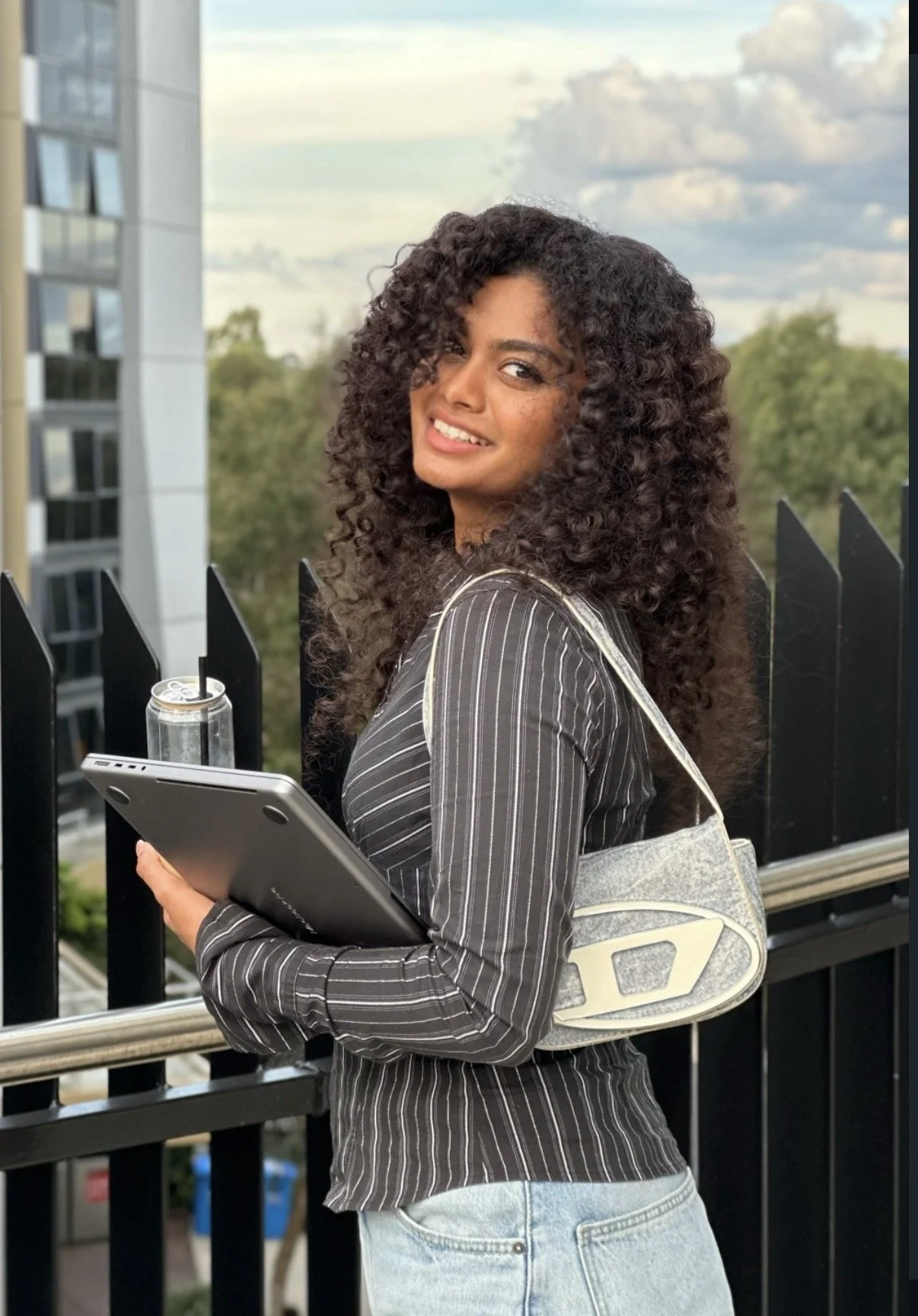 Woman with curly hair holding a tablet and a can with a straw, standing outdoors near a black fence, with buildings and trees in the background.