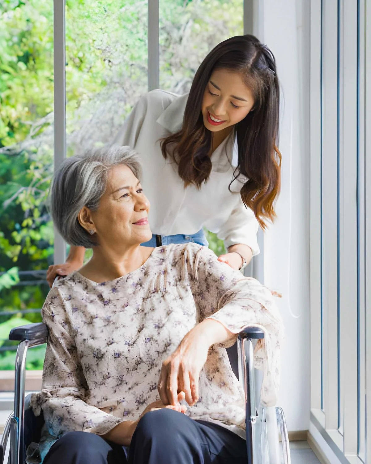 A young woman and an elderly woman in a wheelchair share a happy moment together indoors near large windows with greenery outside.