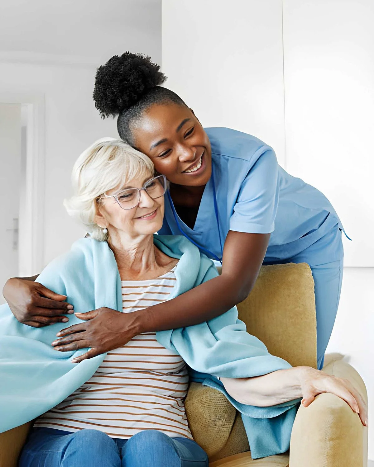 A young healthcare worker hugging an elderly woman sitting on a chair in a medical setting.