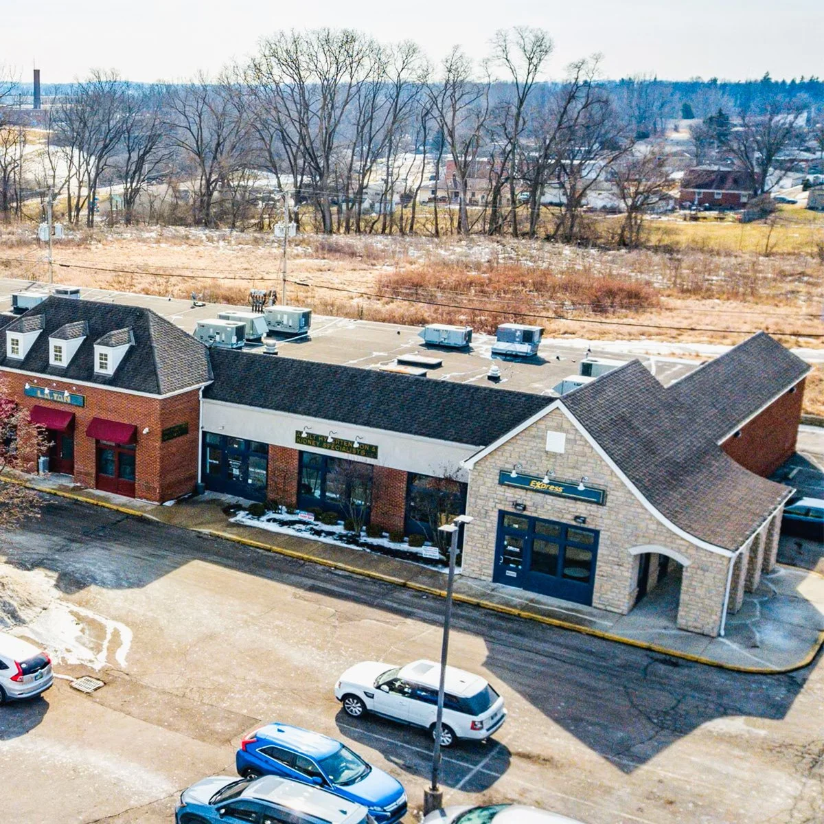 An exterior view of Platinum Circle Clubhouse with three storefronts, including a sign for an insurance agency, a mortgage company, and an express store, with a parking lot and cars in front, and a background of leafless trees and fields.