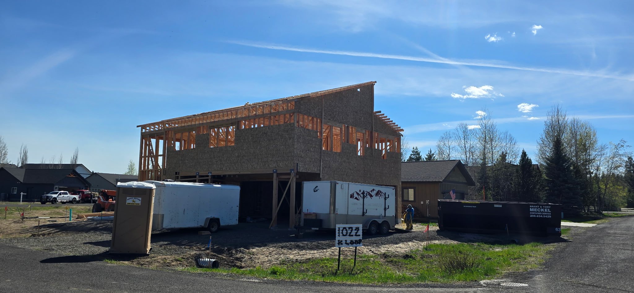A house under construction showing a wooden framework and partially completed exterior walls, with construction trailers and building materials in the foreground, under a blue sky with some clouds.