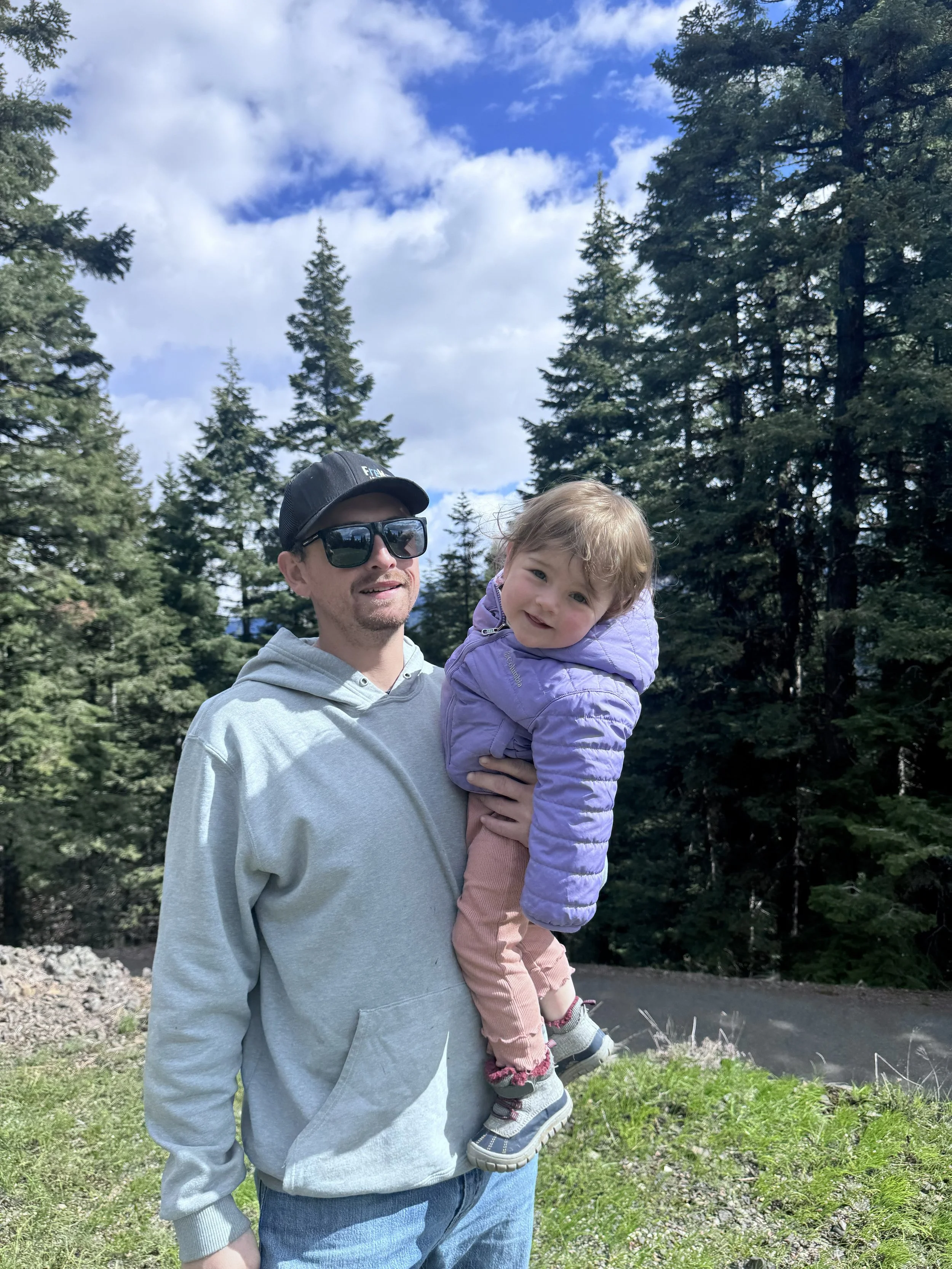 Man wearing sunglasses and a cap holding a young girl in a light purple jacket, outdoors with trees and blue sky in the background.