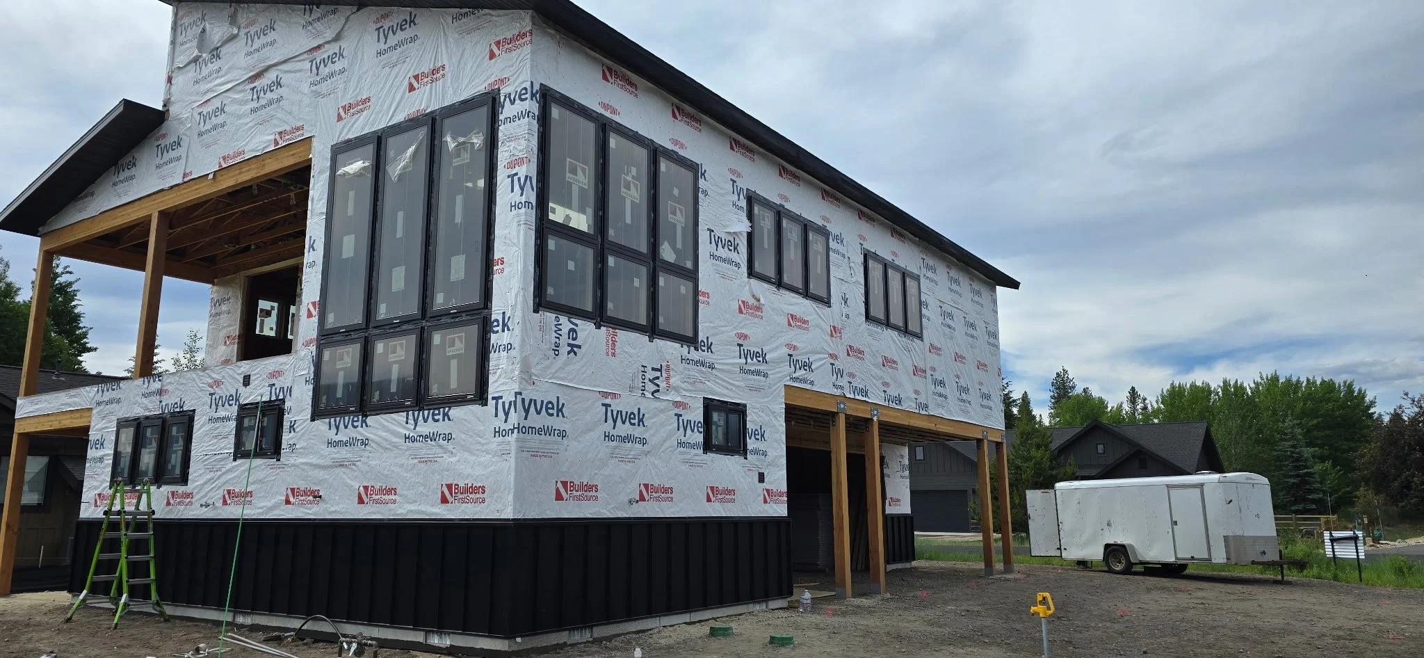 A two-story house under construction with wrapped exterior walls, multiple window frames installed, and an porch area being built. Construction materials and a white trailer are visible on site.