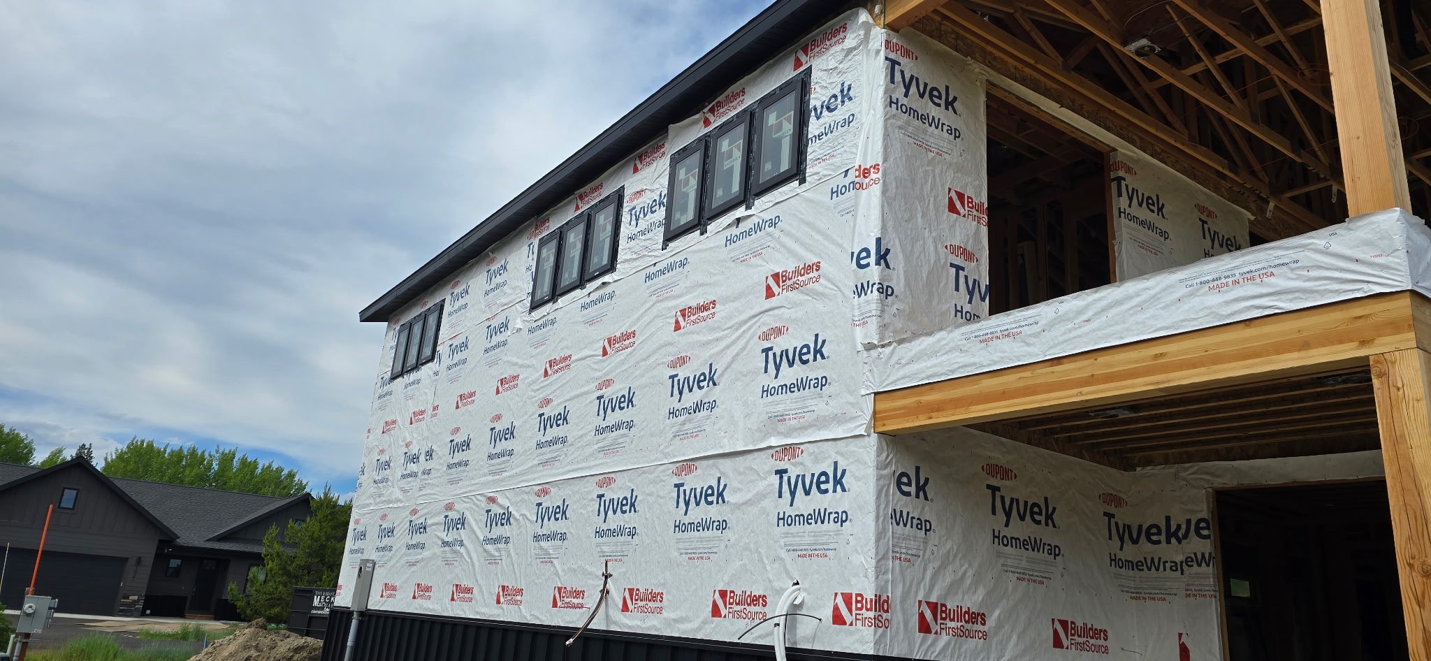 Exterior of a two-story house under construction with weather-resistant wrap and black-framed windows on the upper level, with visible wooden framing underneath the roof and on the right side.