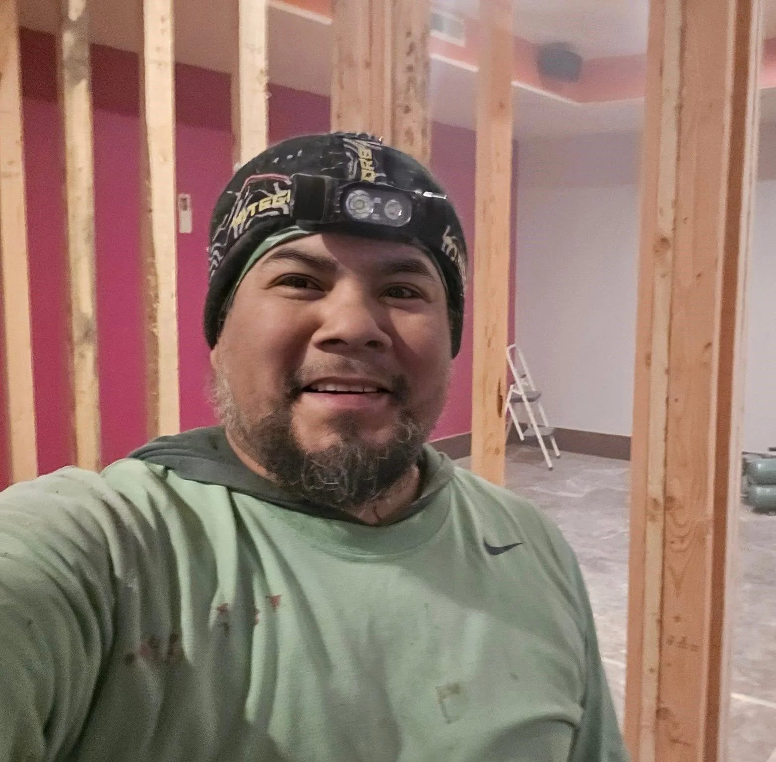 A man wearing a green Nike shirt and a black headlamp takes a selfie inside a building under construction, with exposed wooden framing and a ladder in the background.