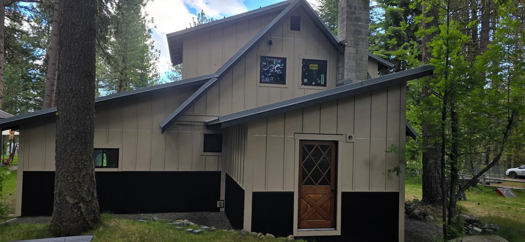 A house in the woods with a sloped roof, beige vertical siding, and a stone chimney, surrounded by tall trees.