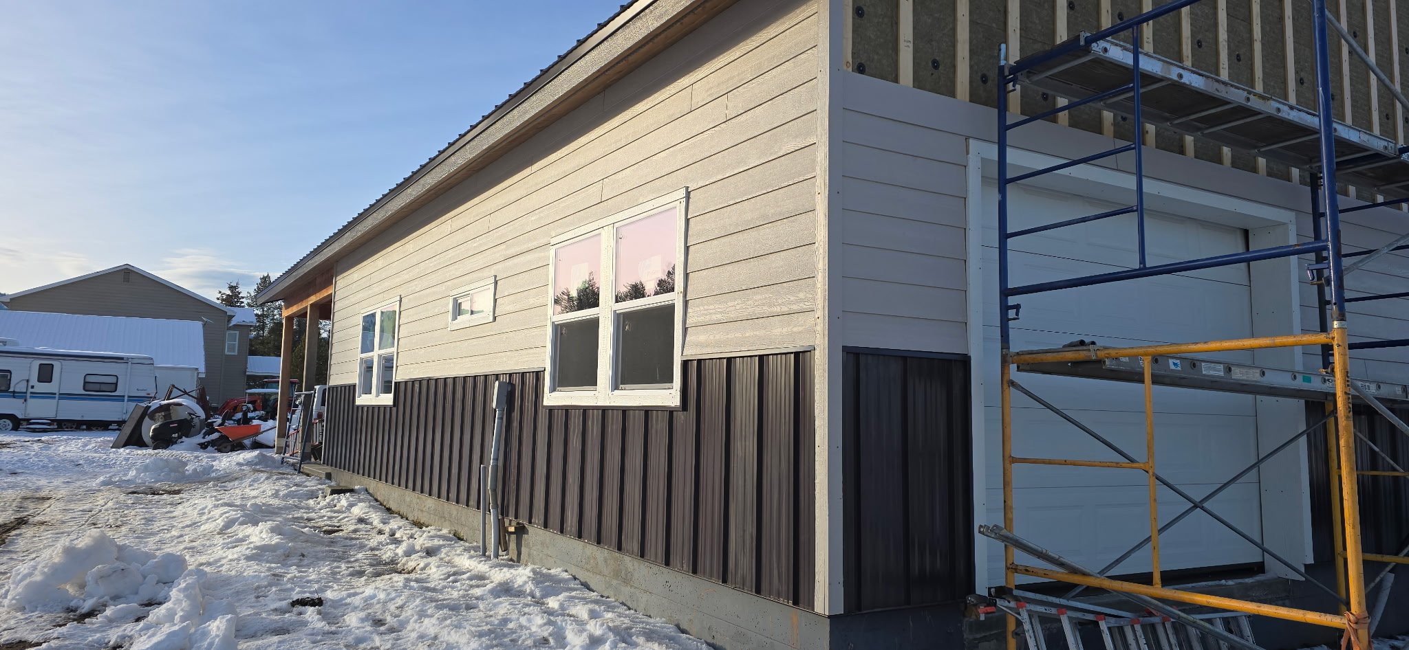 A house under construction with siding partially installed, scaffolding on the side, and snow on the ground.