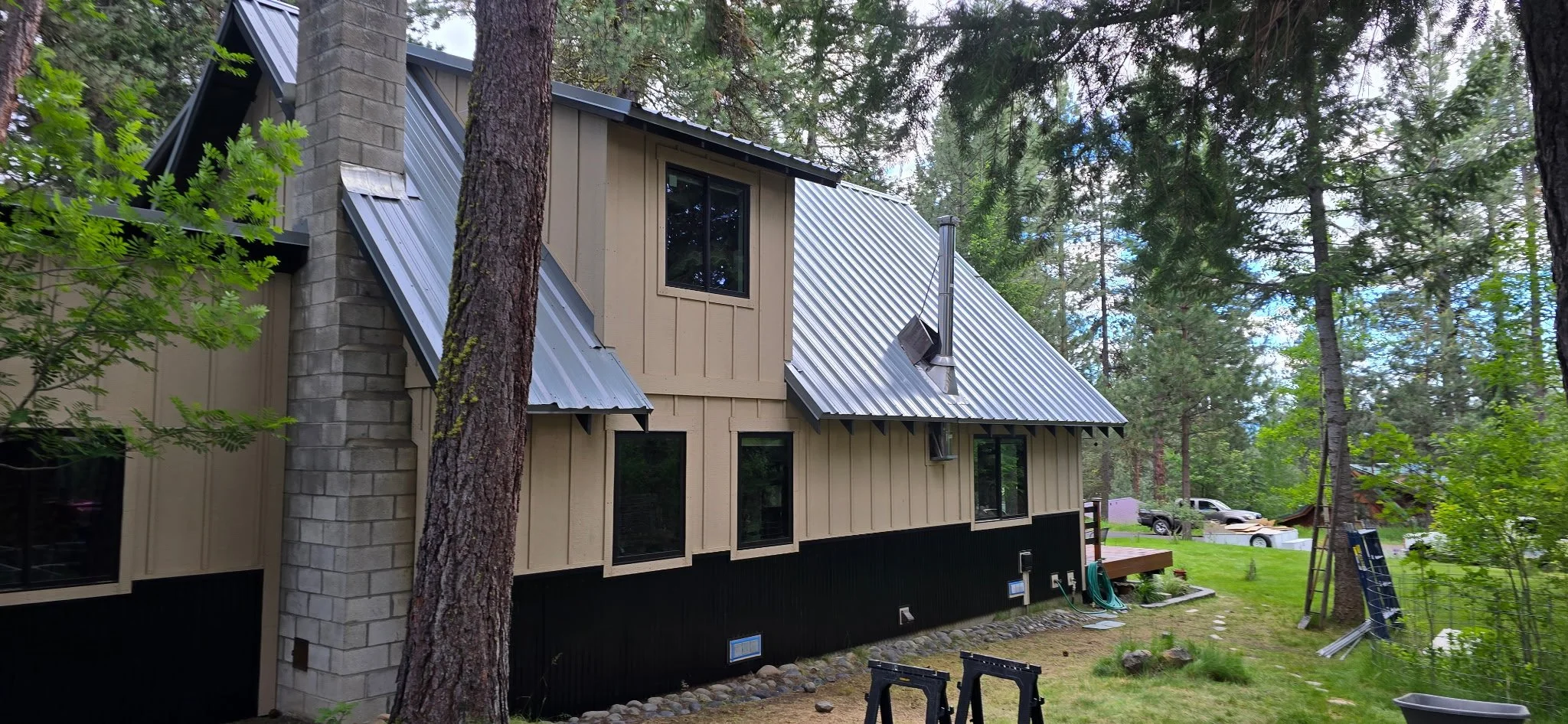 A two-story house with a beige exterior, black bottom section, and metal roof, situated among tall pine trees with a grassy yard and construction tools nearby.