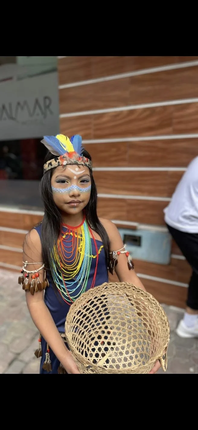 Young Kichwa girl wearing traditional Indigenous clothing, face paint, colorful bead necklaces, and feather adornments while holding a woven basket.