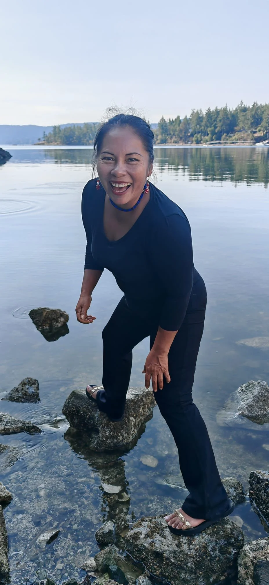 Samay Taki standing on river rocks in a forest river beneath a blue sky.