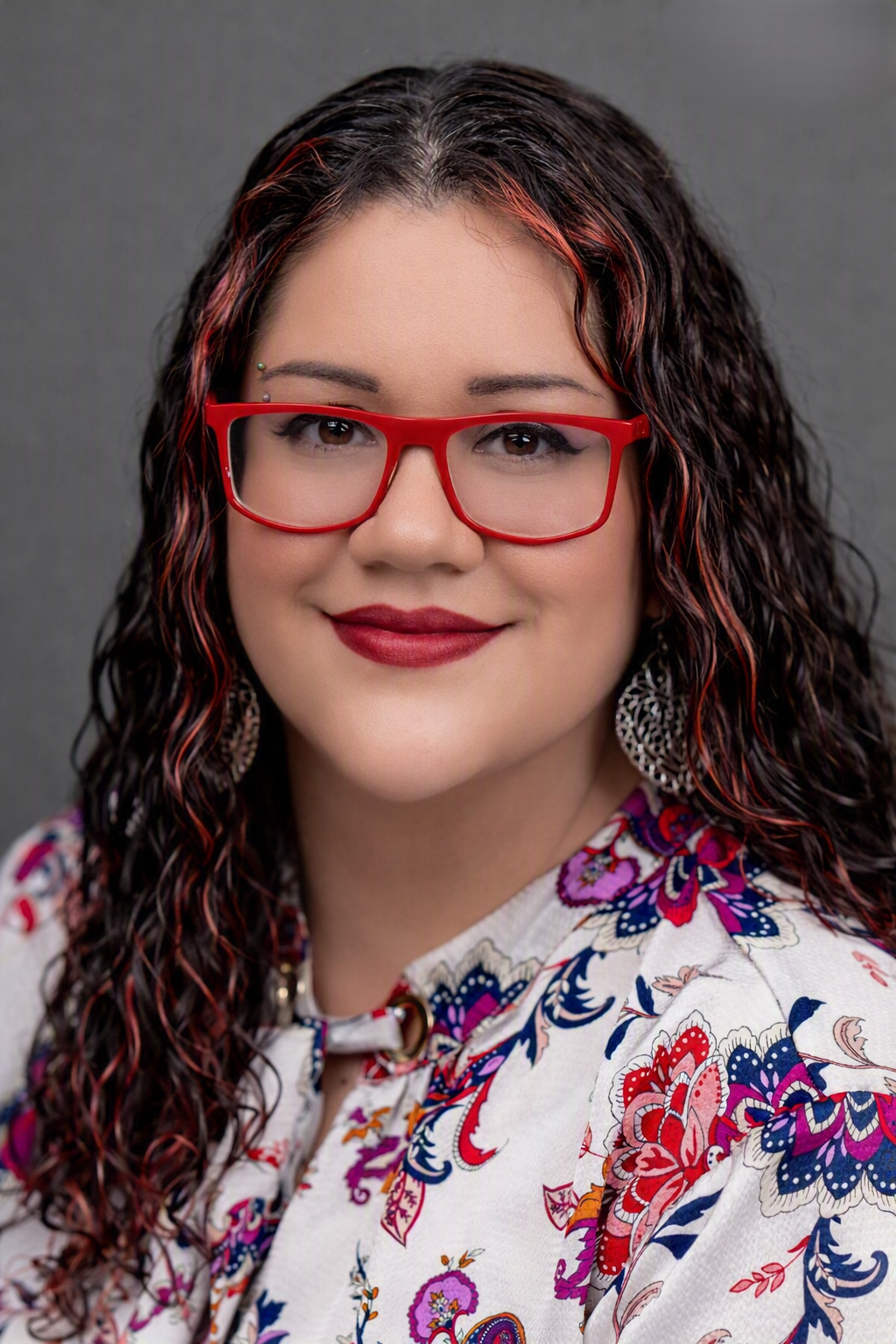 A woman with long wavy dark hair with red highlights, red glasses, wearing a floral patterned top, smiling against a gray background.