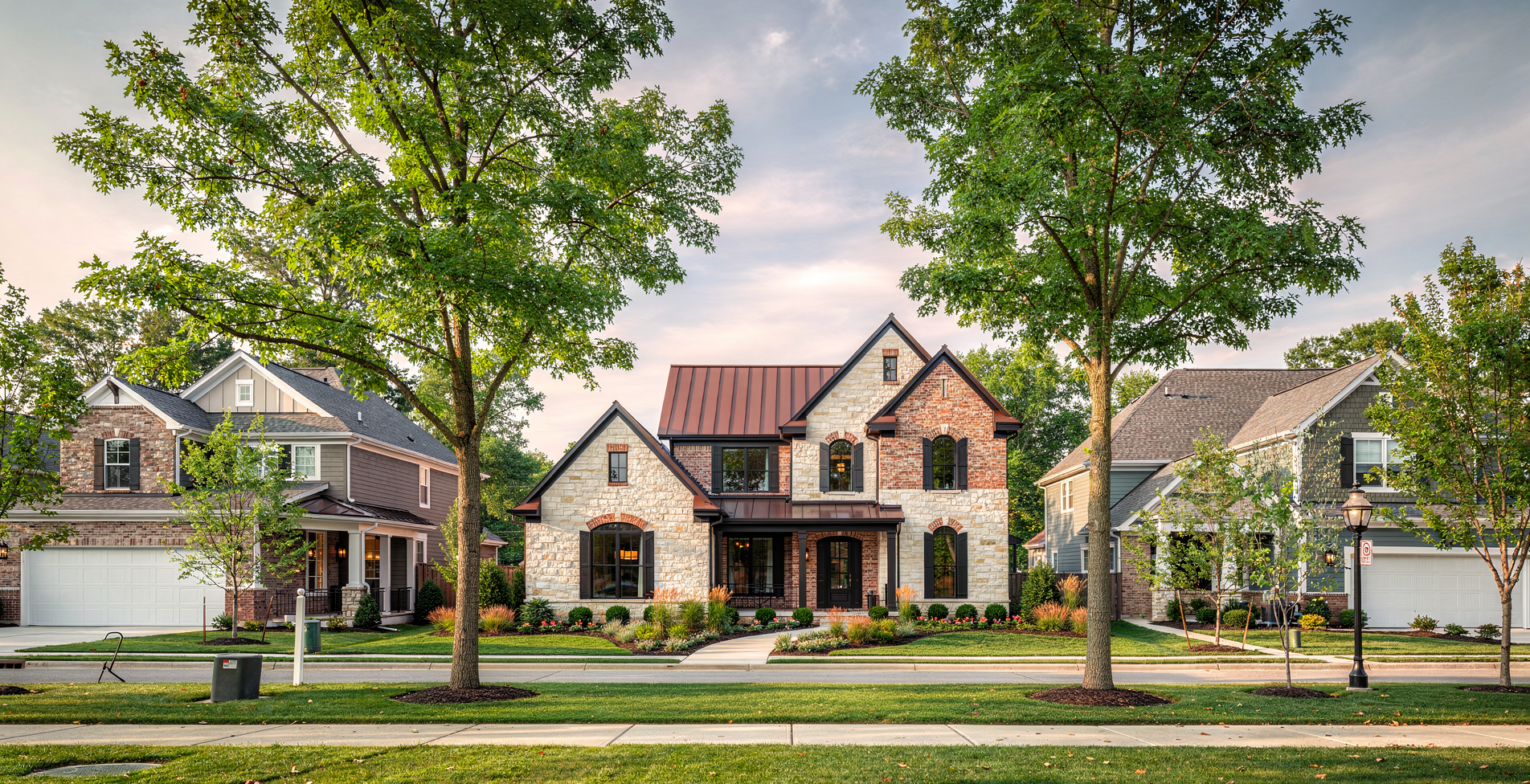 Suburban neighborhood with modern houses, tall trees, well-maintained lawns, a sidewalk, street lamps, and a clear sky at dusk.