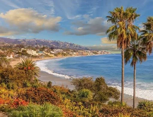 Coastal view of a beach with palm trees, ocean waves, and distant hills under a partly cloudy sky.