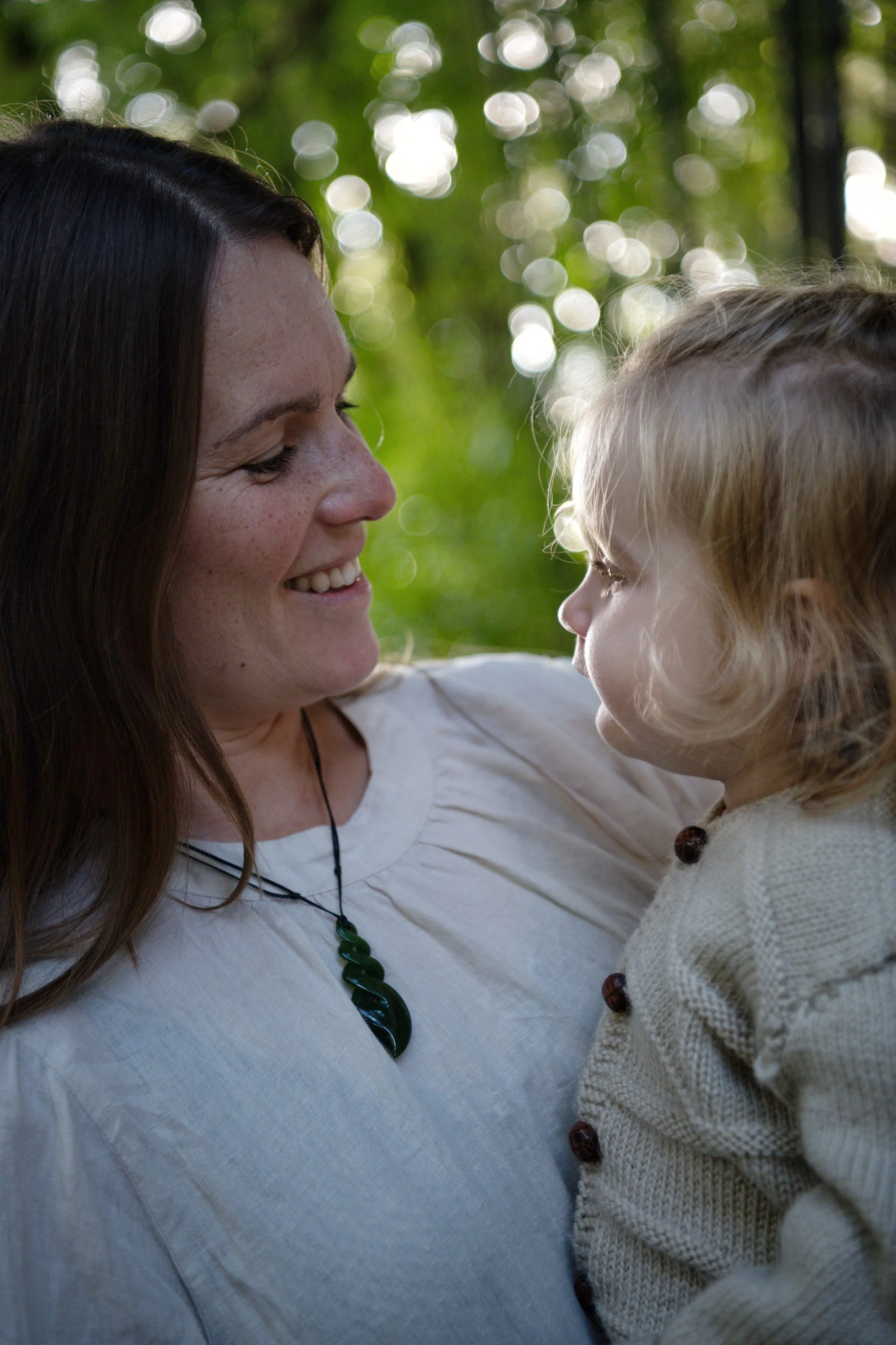 A woman and a young girl facing each other outdoors surrounded by blurred greenery. The woman has long dark hair, freckles, and is smiling warmly. The girl has light hair with curls and is wearing a beige sweater with buttons.