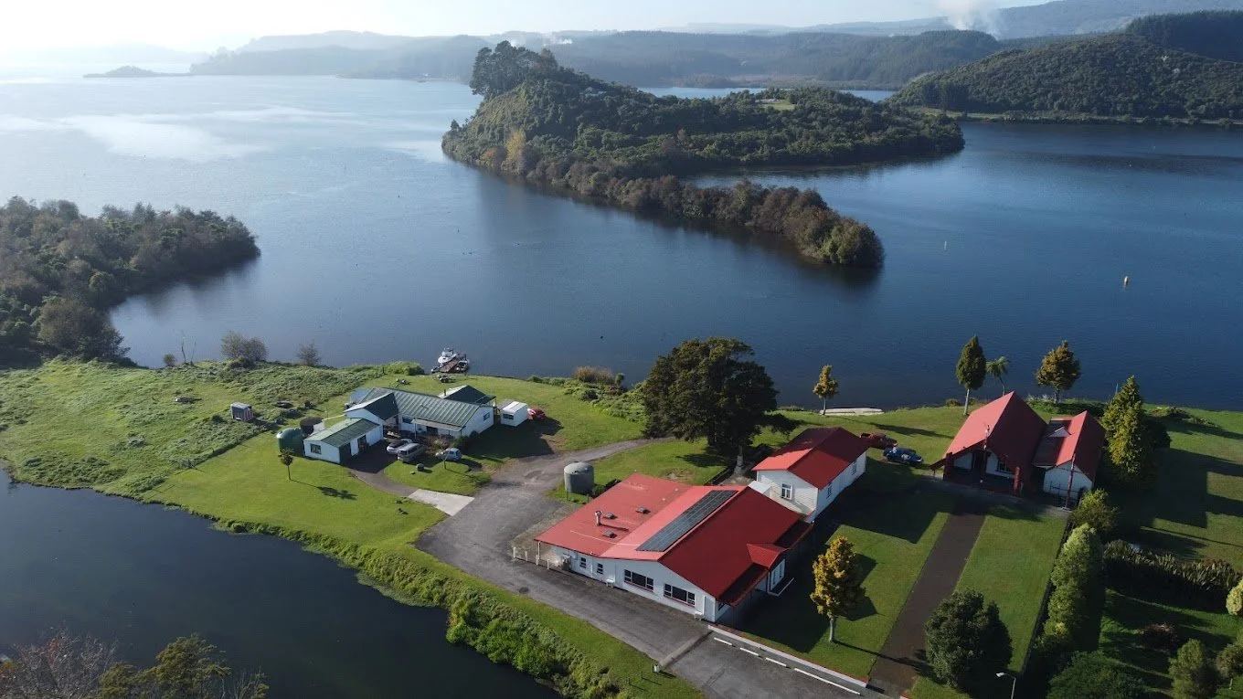Aerial view of a lakeside property with multiple buildings, green lawns, trees, and surrounding water bodies, with hills in the background.