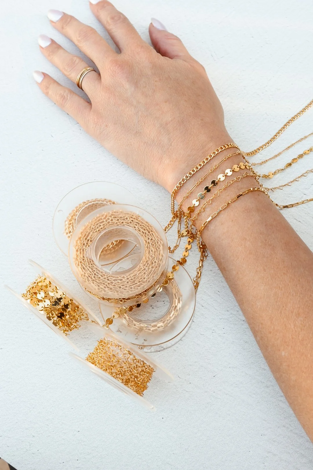A woman's hand with white nail polish and gold rings displays multiple 14k gold filled chains on a white surface, along with rolls of 14k gold filled permanent jewellery chain and accessories.