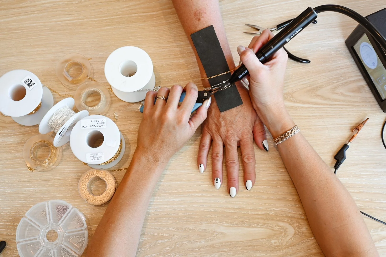 A permanent jewellery artist is welding a bracelet onto a clients arm using pliers; various spools of permanent jewellery 14k gold filled chains, and jewellery tools are spread on the wooden table.