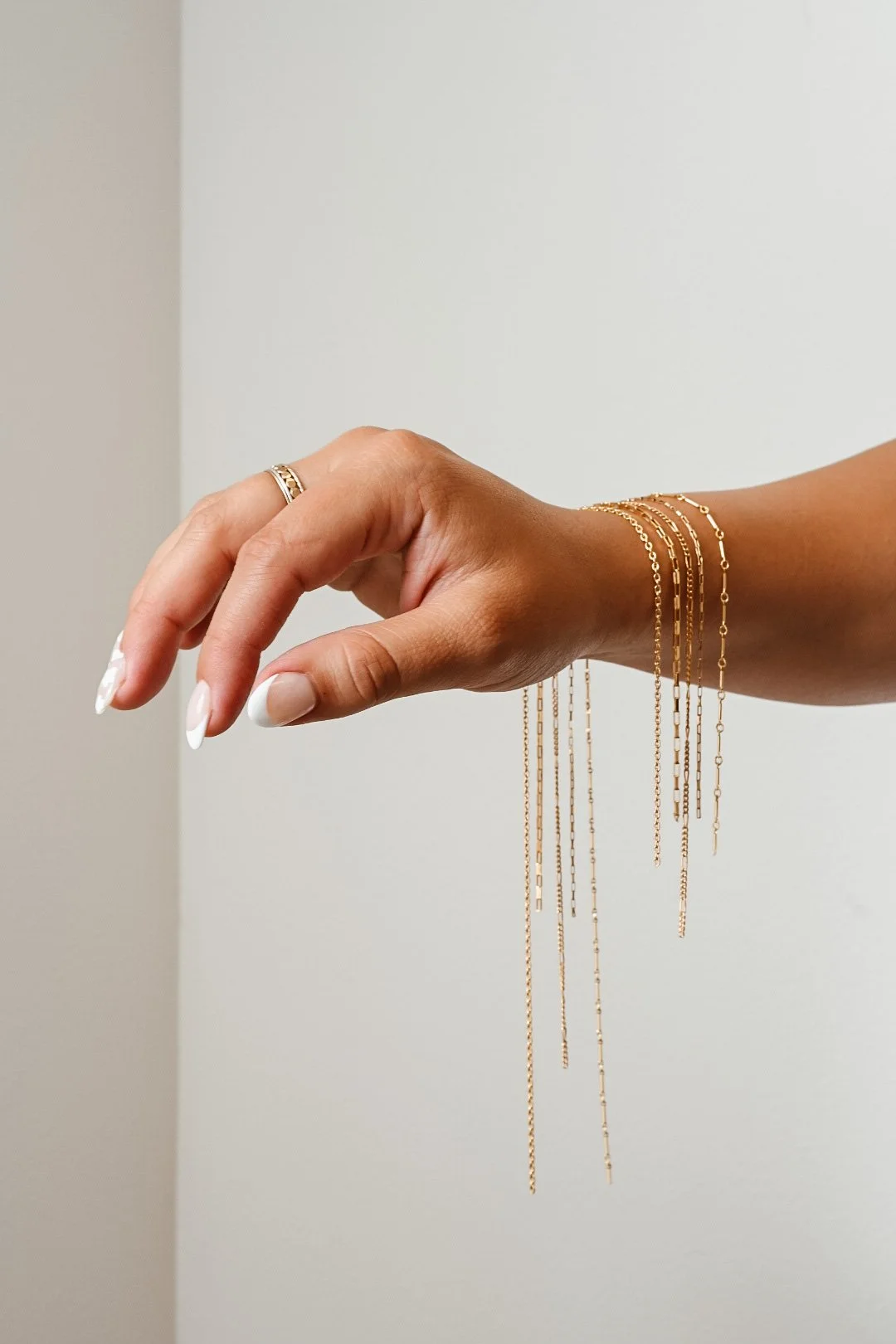 A woman's hand with a gold ring and several 14k gold filled chains for permanent jewellery hanging from her wrist against a plain wall.