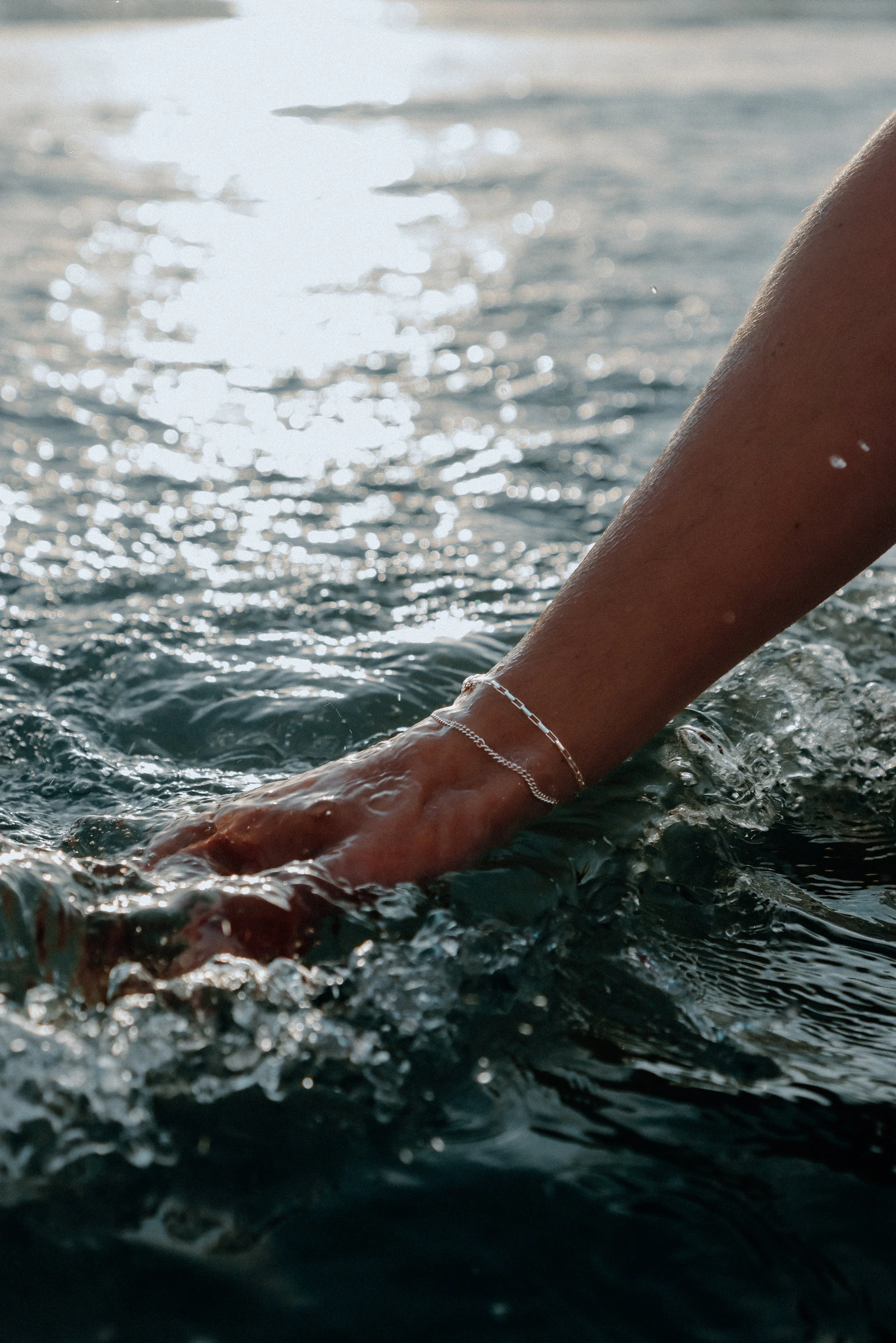 Close-up of a person's arm submerged in water, wearing a delicate sterling silver permanent bracelet, with sunlight reflecting off the water's surface.