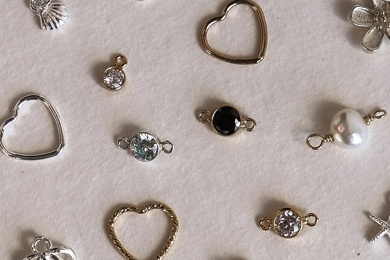 Various permanent jewellery connectors and charms, including heart-shaped and flower-shaped pieces, with some featuring rhinestones, pearls, and different metal finishes, placed on a light-coloured background.