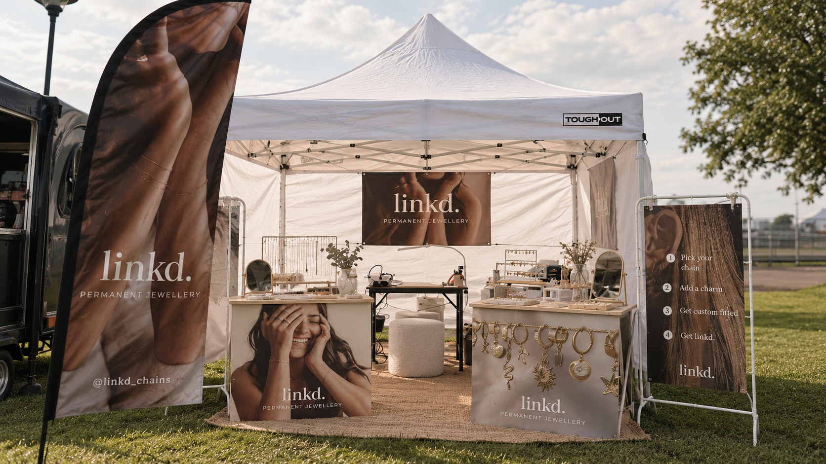 Outdoor market permanent jewellery gazebo with banners and display tables featuring links to permanent jewellery collection, gold rings and charms, under a white gazebo with clear sky.