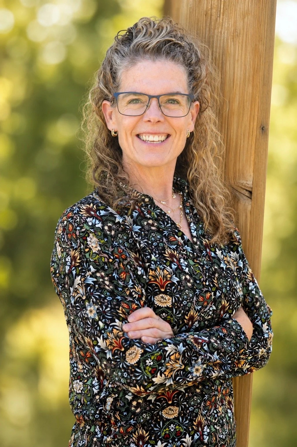 A woman with curly hair, glasses, and earrings is smiling outdoors, standing next to a wooden post, wearing a colorful floral-patterned blouse.
