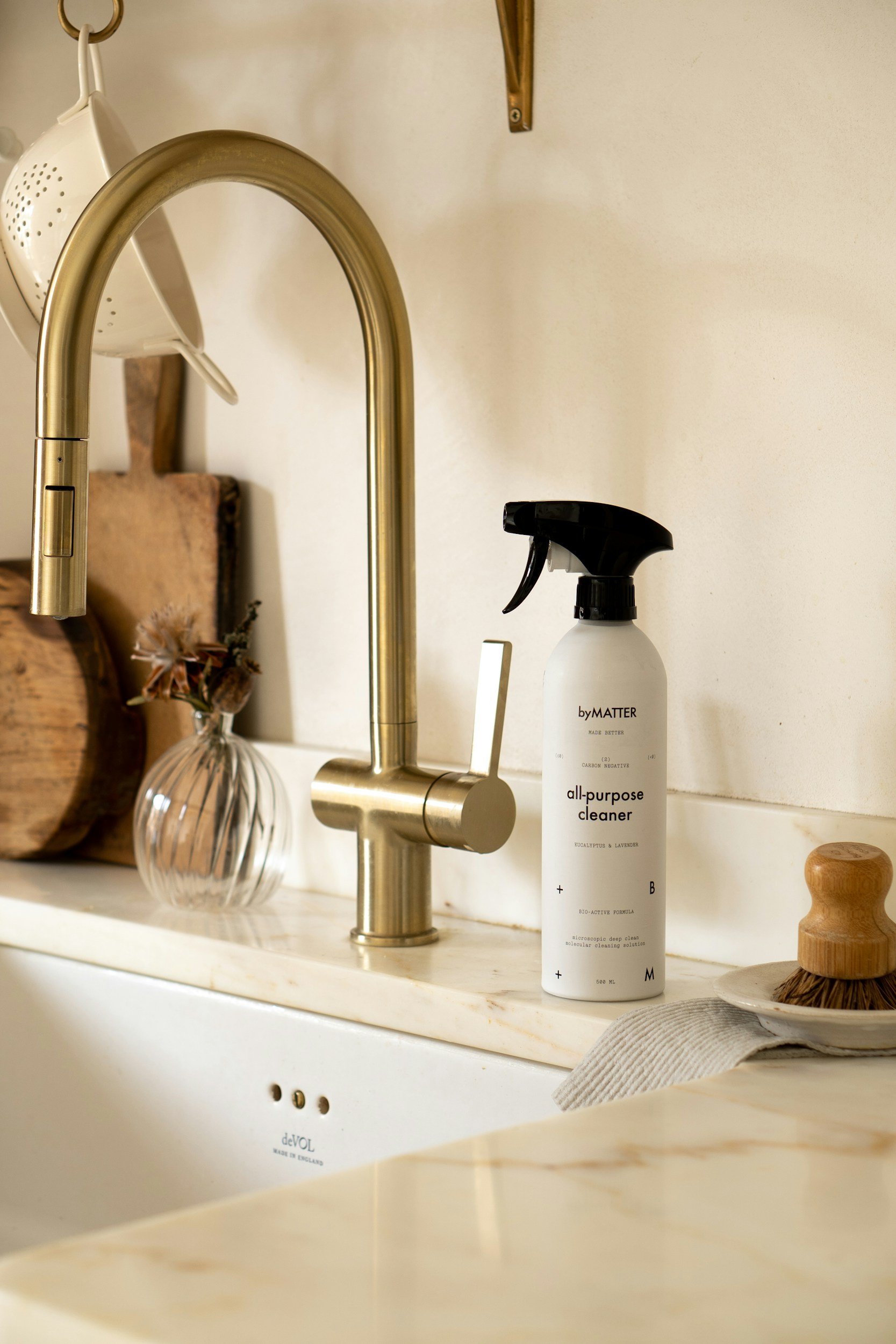 Close-up of a kitchen sink with a gold faucet, a black spray bottle labeled "all-purpose cleaner", a white container labeled "deVOL", a small clear glass vase with dried flowers, a wooden soap dish, a light-colored dish towel, and various kitchen items in the background.