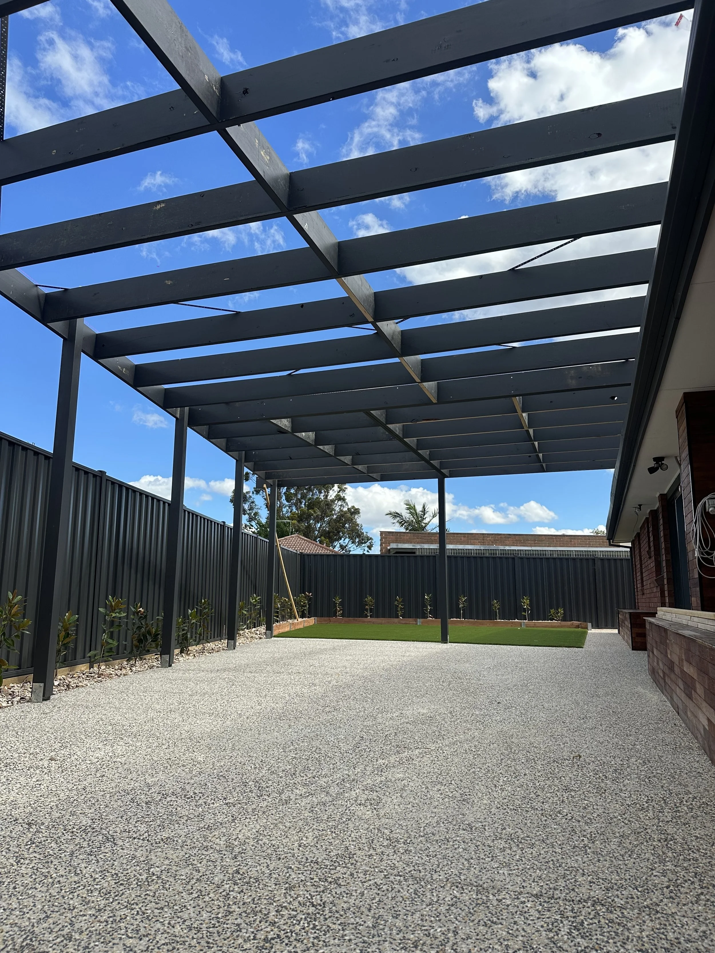 A backyard with a gravel ground, small shrubs along a black fence, a patch of green grass, and a partially constructed black pergola against a blue sky with white clouds.