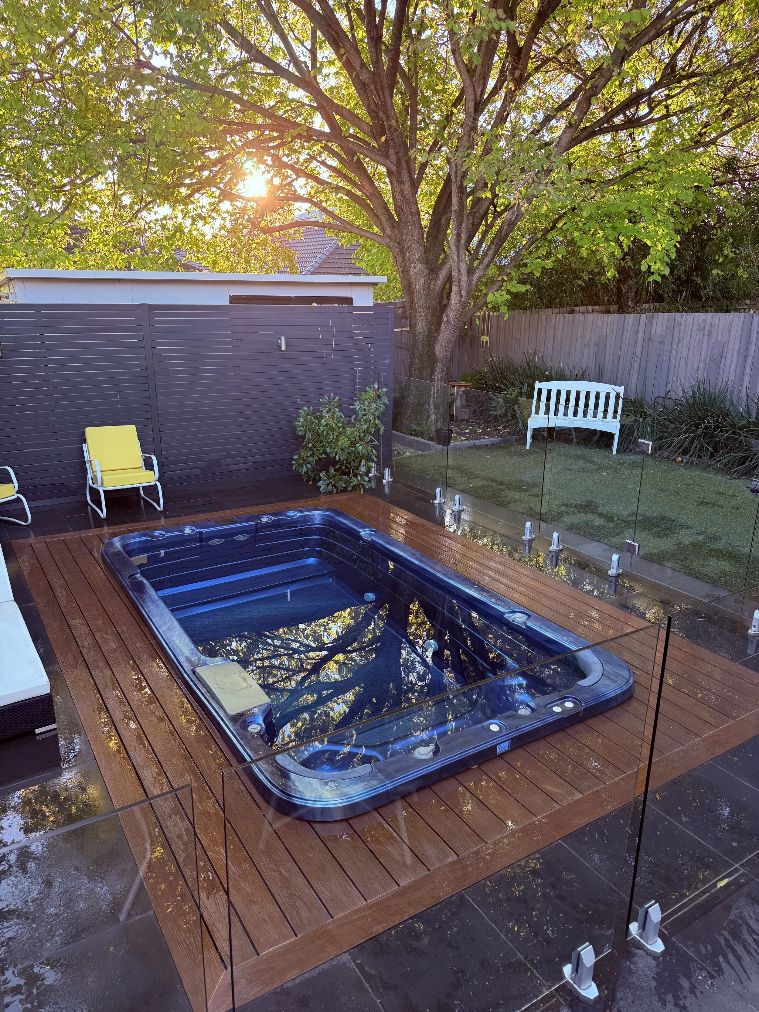 An outdoor backyard with a hot tub on a wooden deck, a yellow chair, a white bench, and a large tree with green leaves. The sun is shining through the leaves, creating a bright and inviting atmosphere.