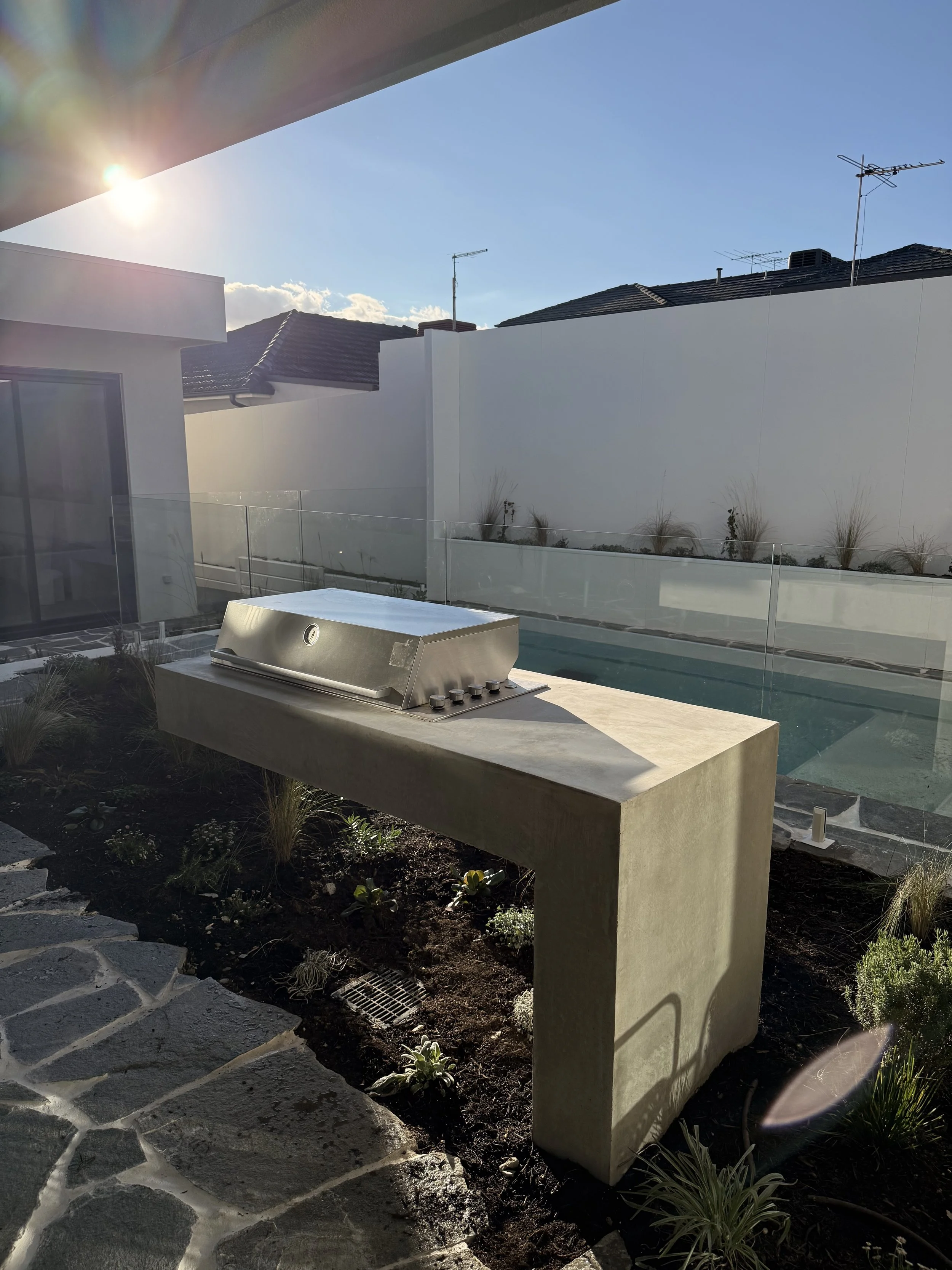 Modern backyard with built-in stainless steel grill on a stone countertop, small landscaped garden with plants and dirt, a tall beige concrete bench, and a white wall surrounding the area under a sunny sky.