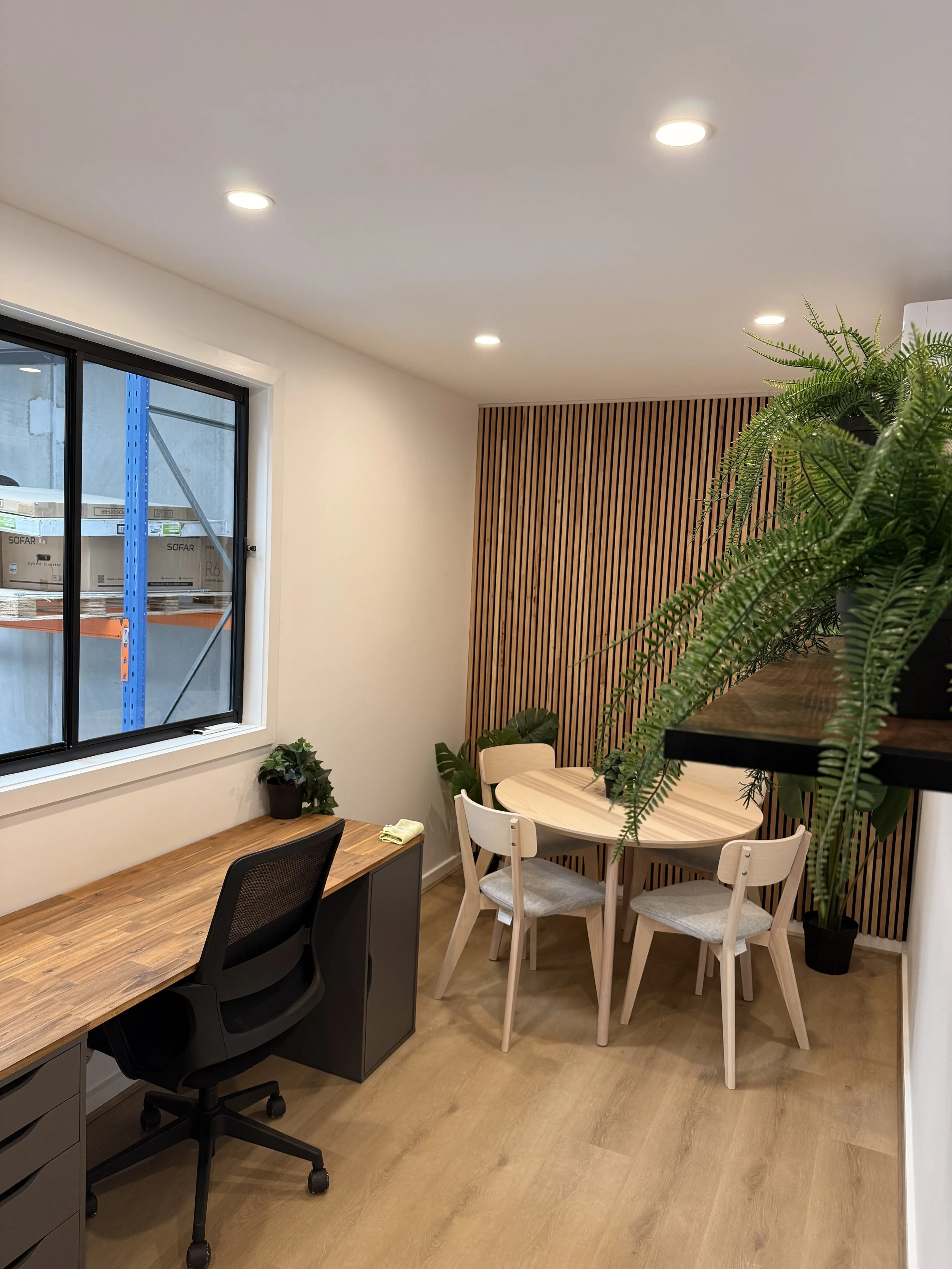 Interior of a modern office space with a wooden table, chairs, plants, and a window showing industrial shelving outside.