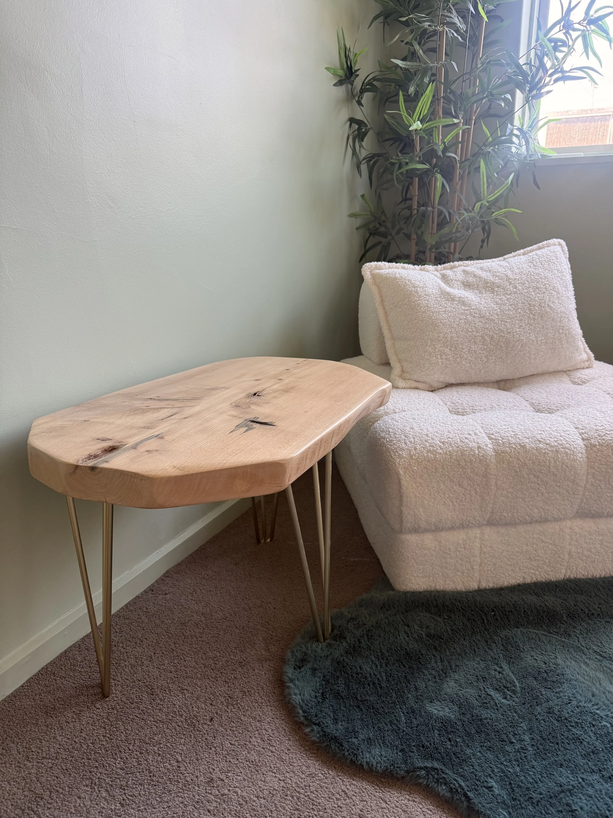 A wooden side table with metal legs positioned beside a white textured sofa with a plush cushion. In the background, a large potted plant with green leaves is near a window, which is allowing sunlight into the room. The floor is carpeted in a pinkish-brown color, and there's a dark gray shaggy rug partially under the sofa and table.