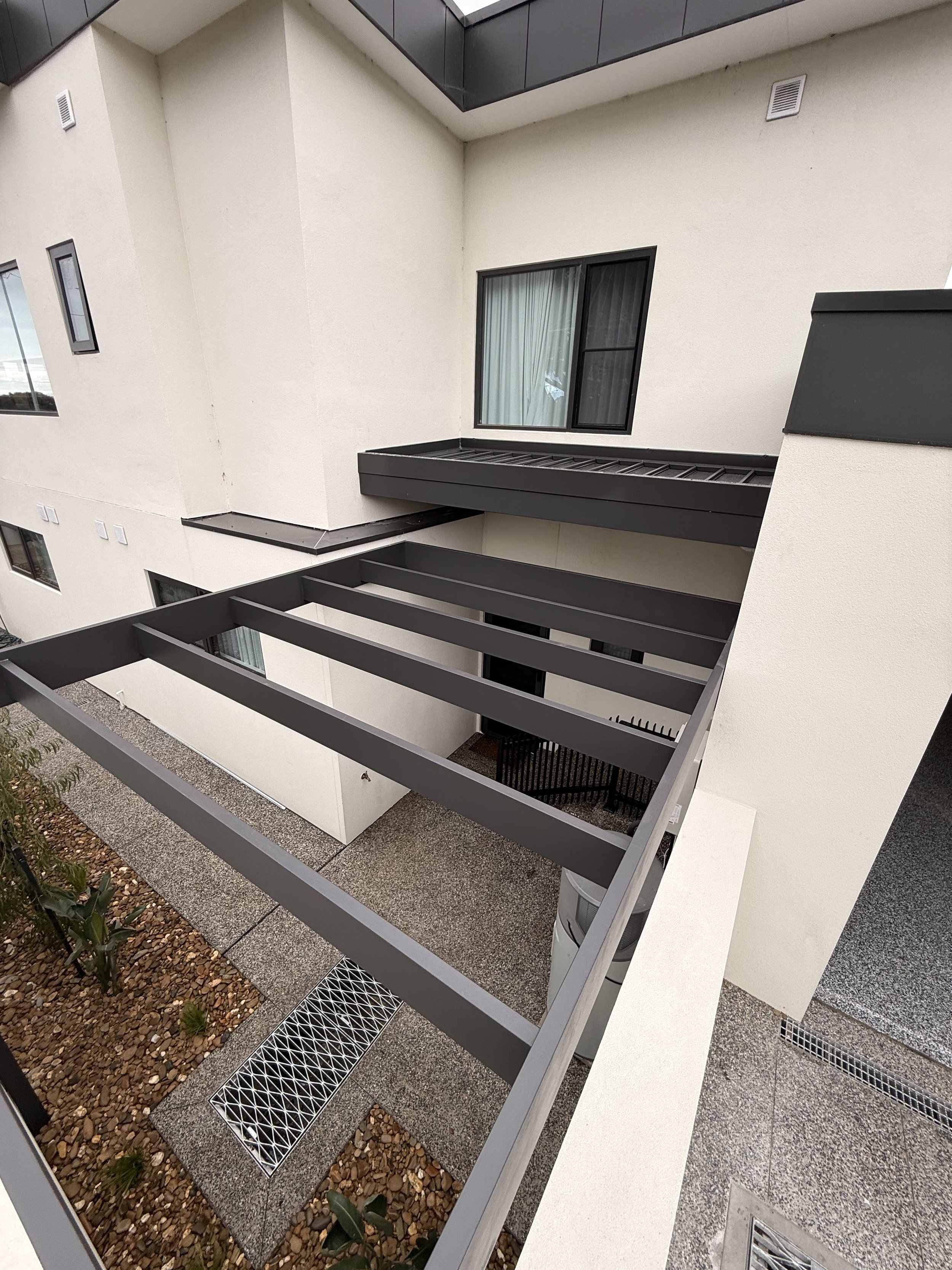 View of an apartment building balcony, showing black metal railings, a window with curtains, and a beige exterior wall with vents. The balcony has a concrete floor with gravel landscaping below.