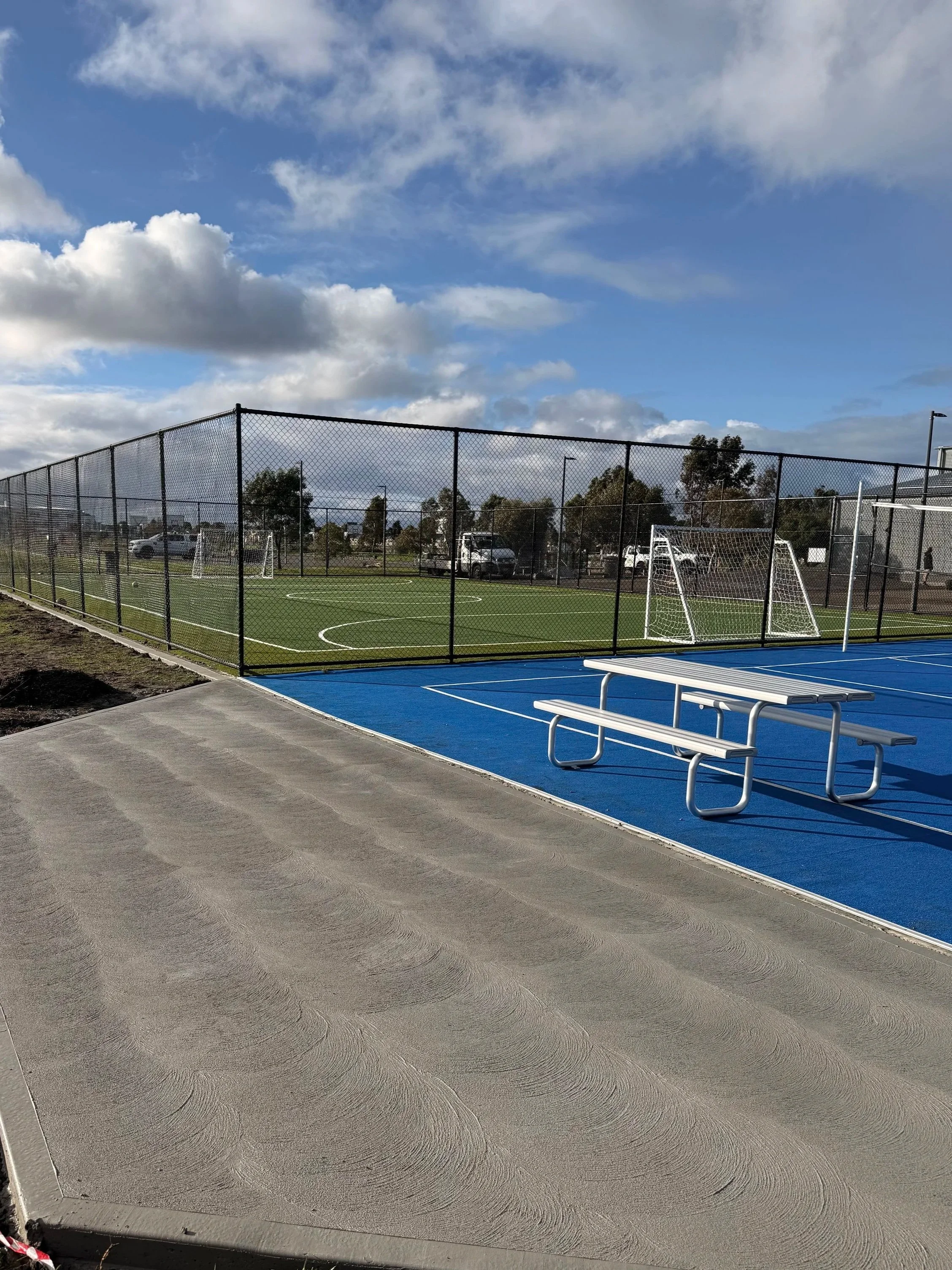 An outdoor sports field with a black chain-link fence, small soccer goals, and a blue section on the ground for seating. The field is surrounded by trees, parked cars, and a cloudy sky.