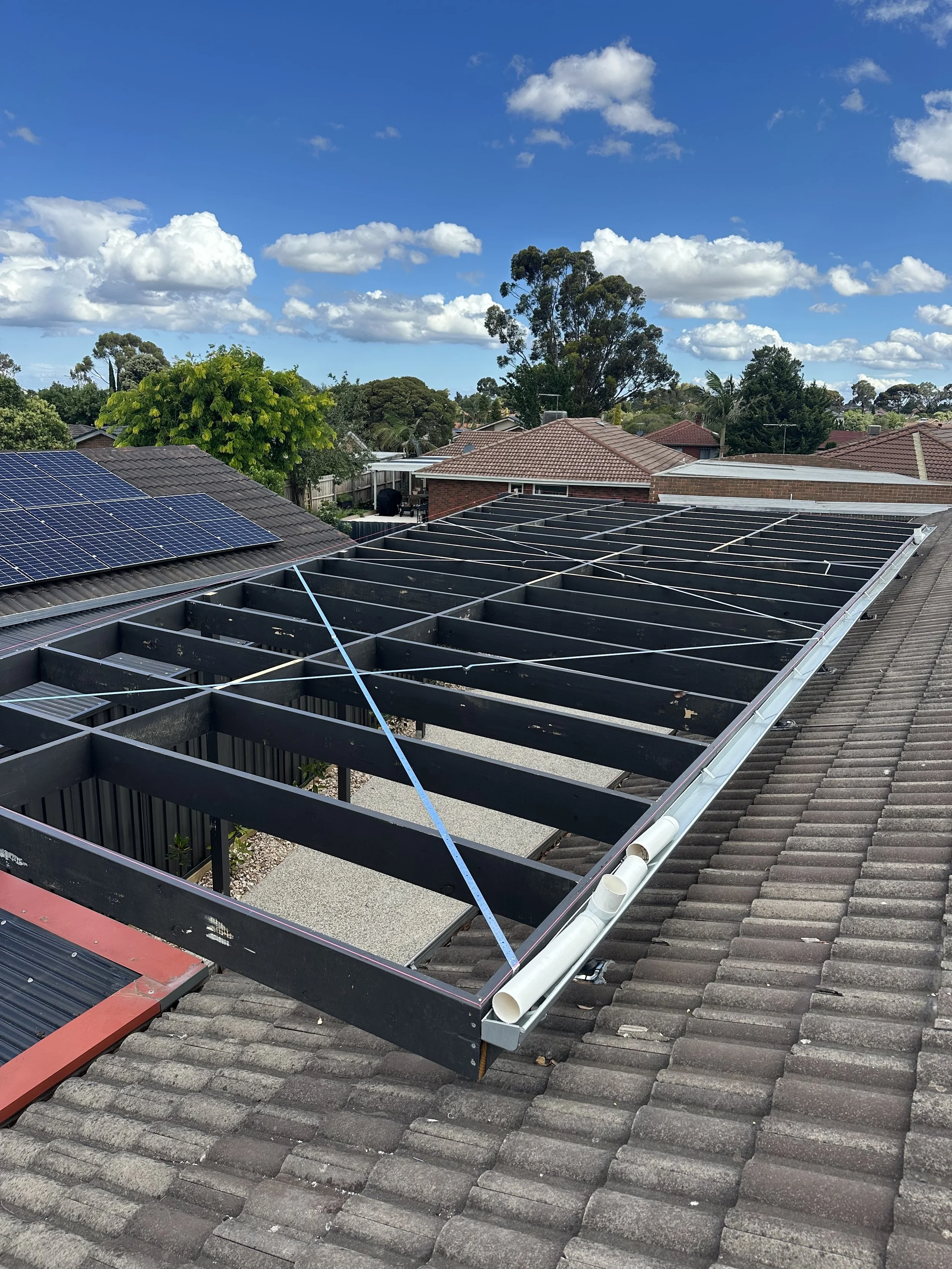 Roof with solar panels being installed, showing the framework and some pipes on the tiles, under a bright blue sky with clouds and trees in the background.