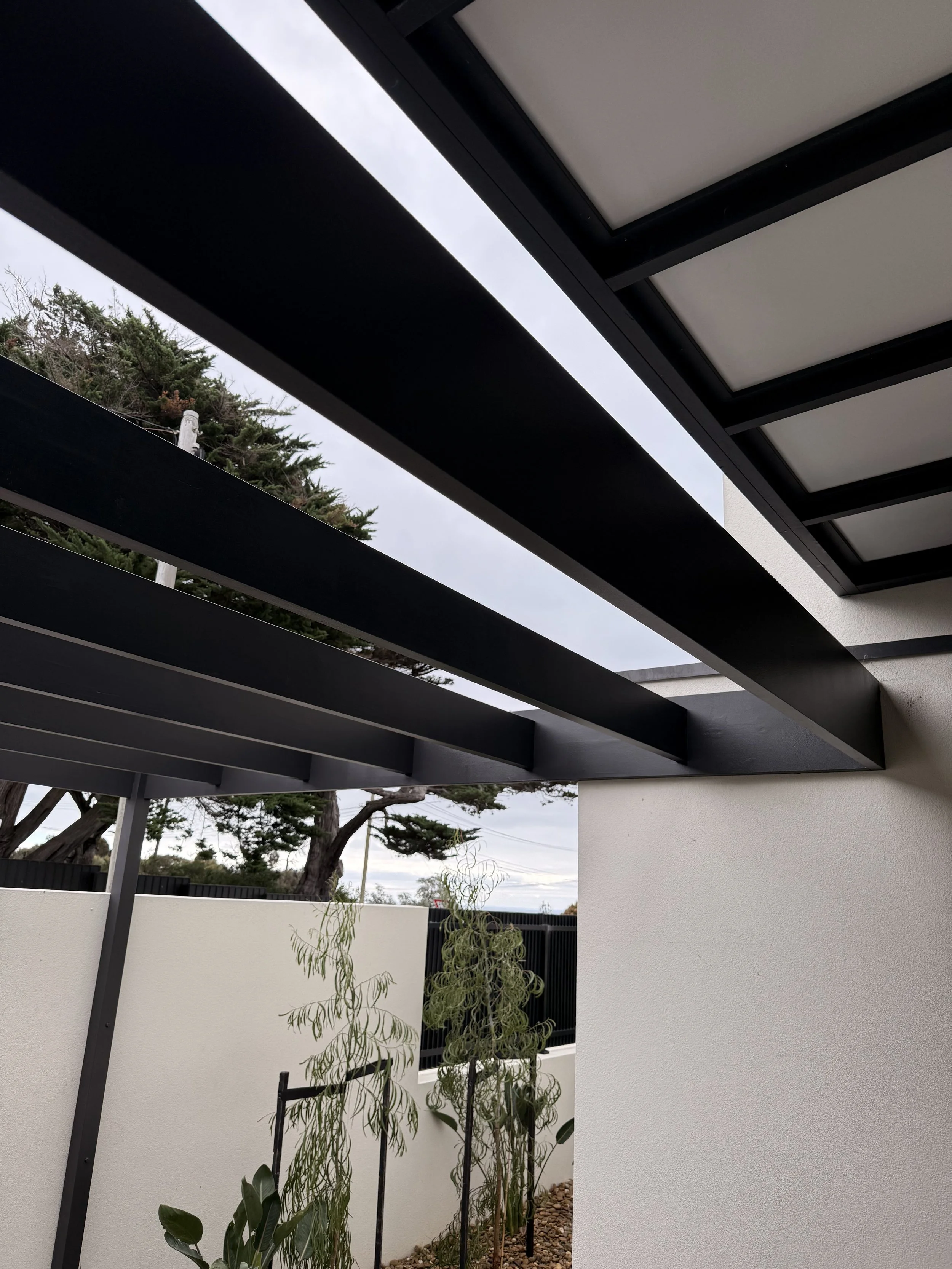 Outdoor patio area with a modern pergola structure featuring black beams and large glass windows on the ceiling. The space is partially enclosed with light-colored walls, with some trees visible in the background, and a few plants in the courtyard.