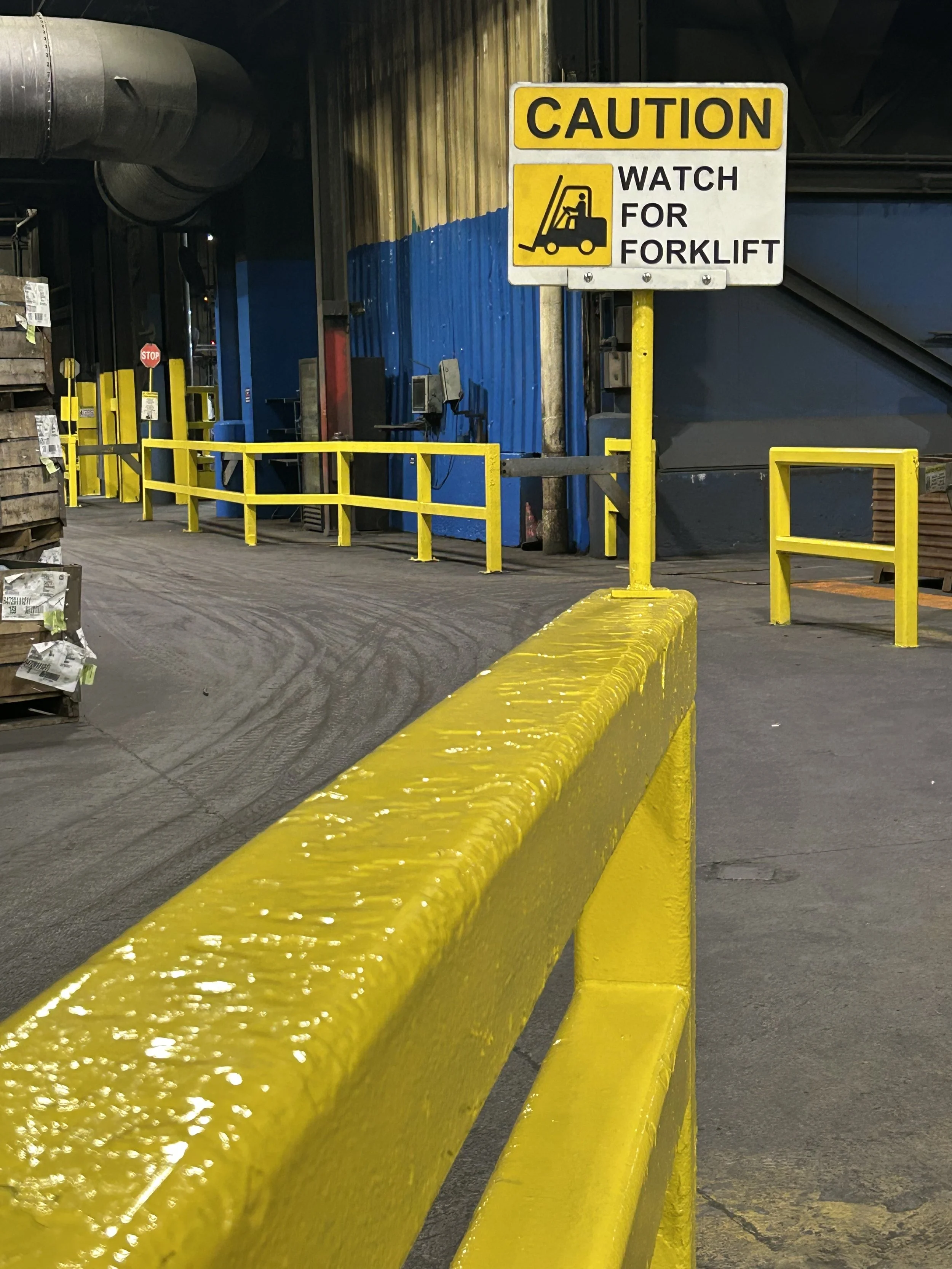 A warning sign inside a warehouse or industrial facility that says, 'Caution: Watch for Forklift,' with a graphic of a forklift, yellow barriers, and industrial equipment and products in the background.