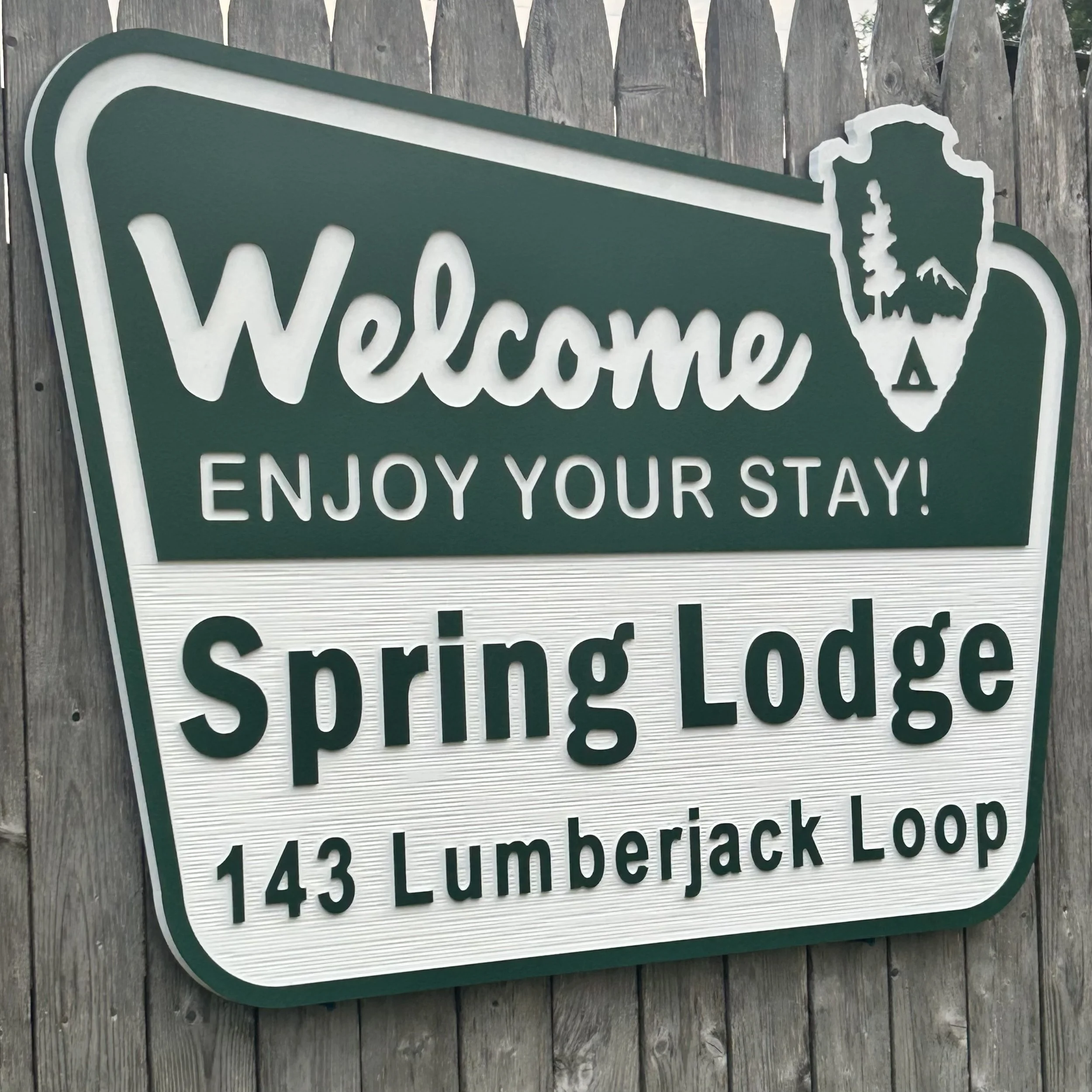 A green and white welcome street sign with a bear outline and mountain image, mounted on a wooden fence.