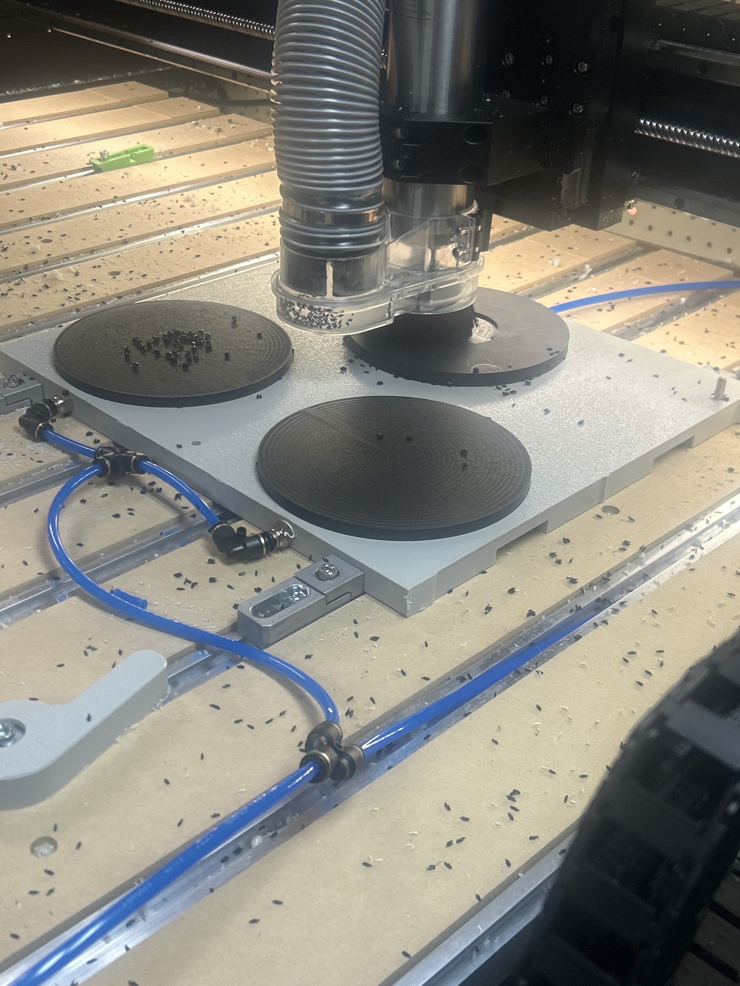 Close-up of a CNC machine cutting black plastic or rubber material, with blue air or coolant tubes, metal grid table, and black plastic or rubber scraps scattered around.