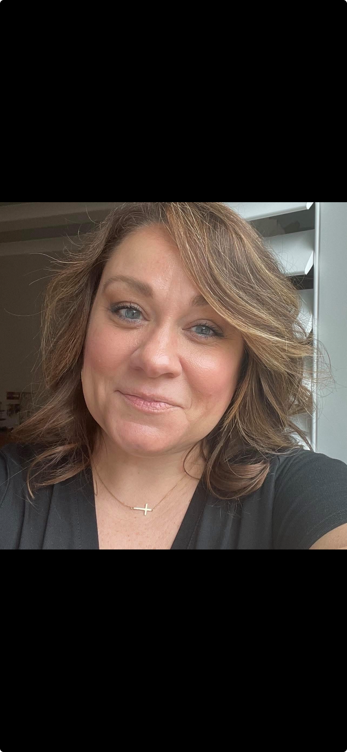 A woman with shoulder-length wavy brown hair and blue eyes takes a selfie indoors. She is wearing a black top, a delicate gold necklace with a cross pendant, and has a subtle makeup look.