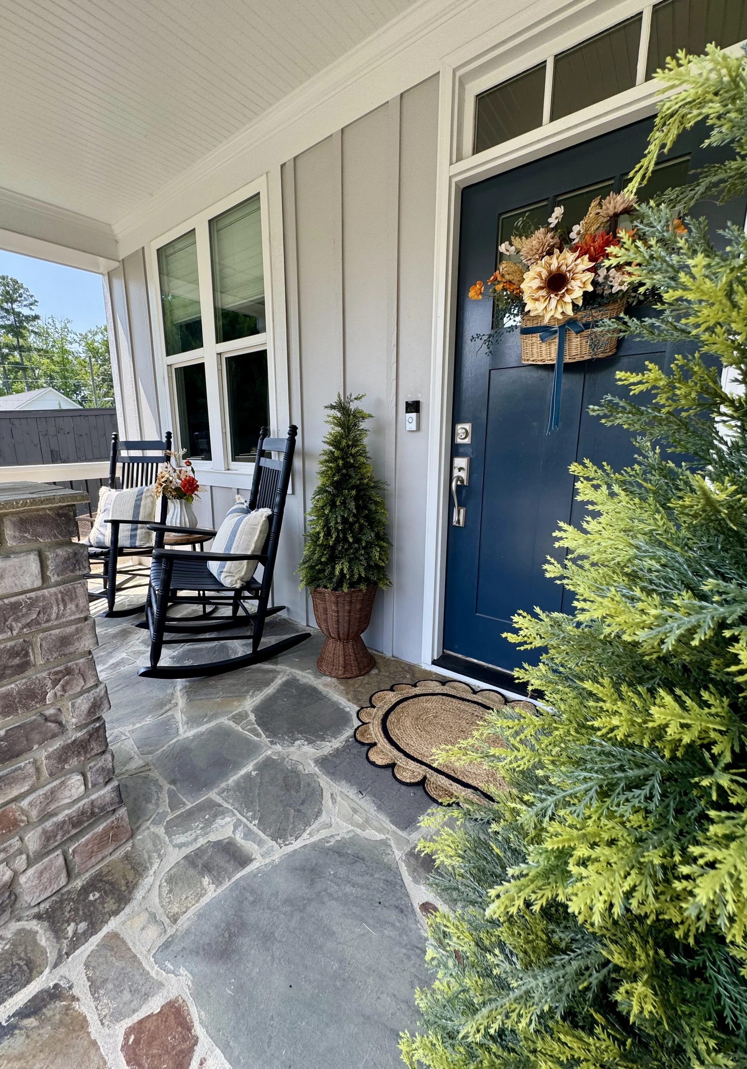 Front porch with two black rocking chairs, a small potted evergreen tree, a colorful fall basket wreath on a blue front door, and a doormat. Part of the house exterior and clear blue sky are visible.