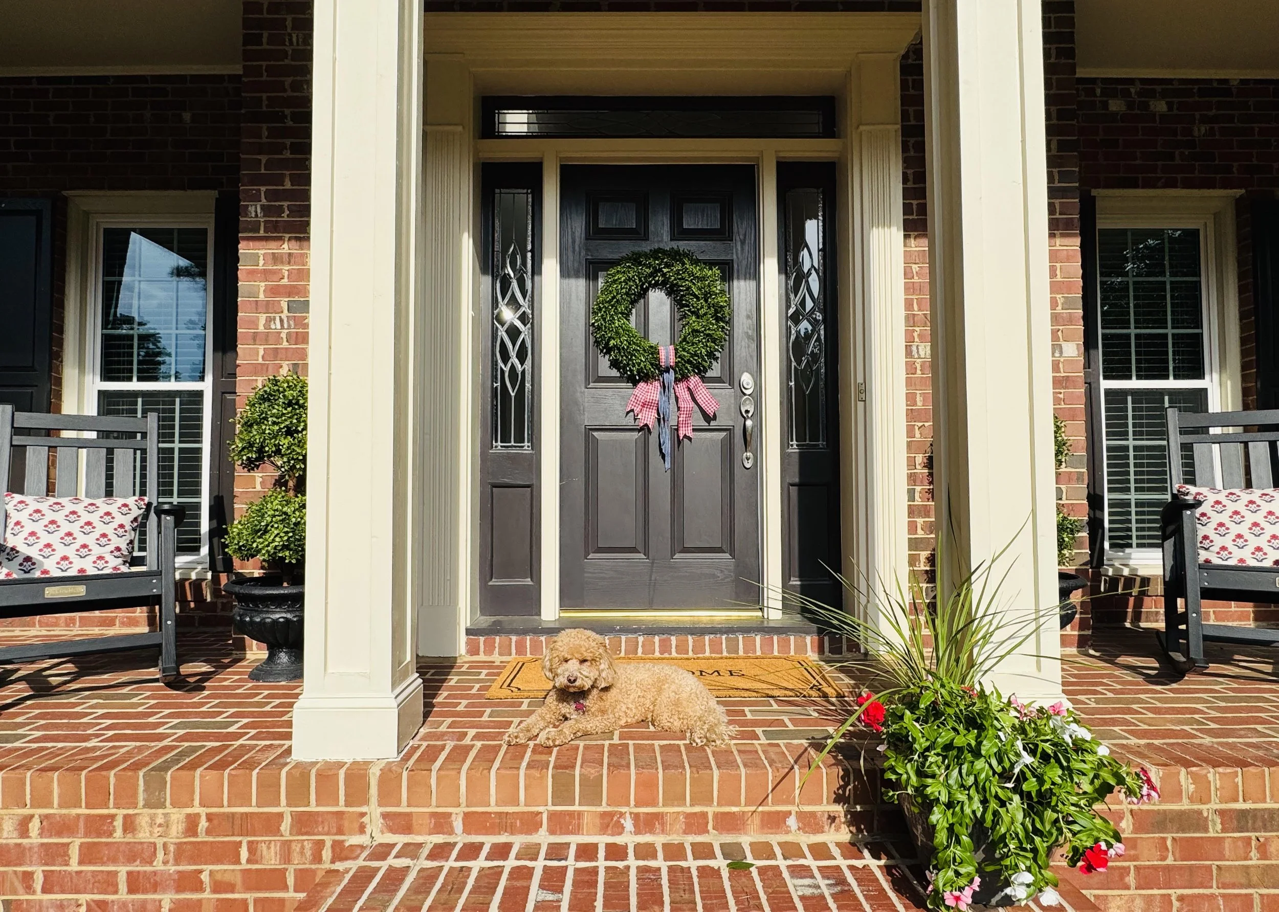 Front porch of a house with brick steps, a black front door decorated with a green wreath tied with red and white ribbons, and two windows on either side. There are two benches with floral cushions, potted plants, flowers, and a small curly-haired dog lying on the brick floor.