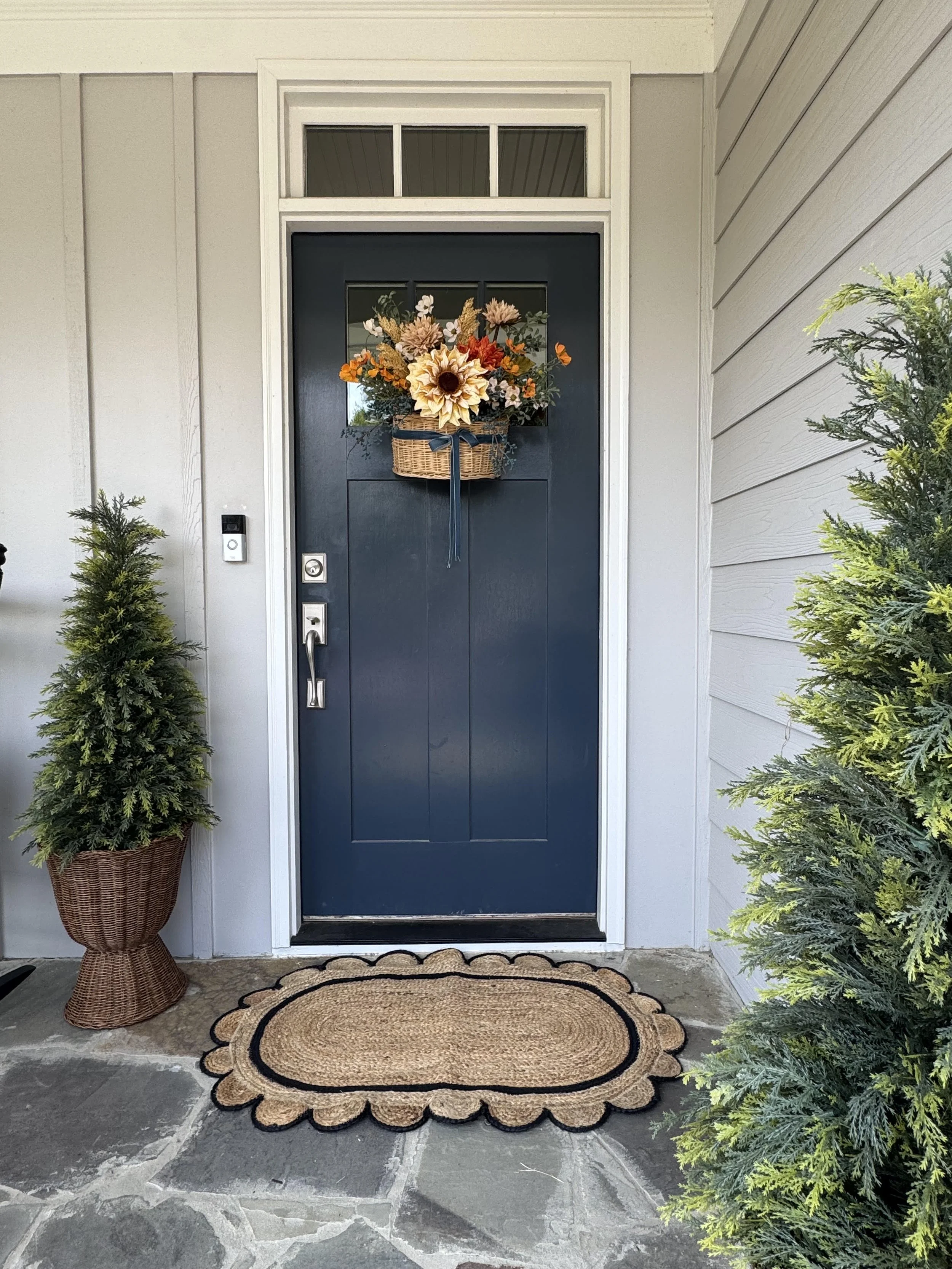Front door with a floral wreath, two potted evergreen plants on either side, a doormat, and gray stone porch floor.