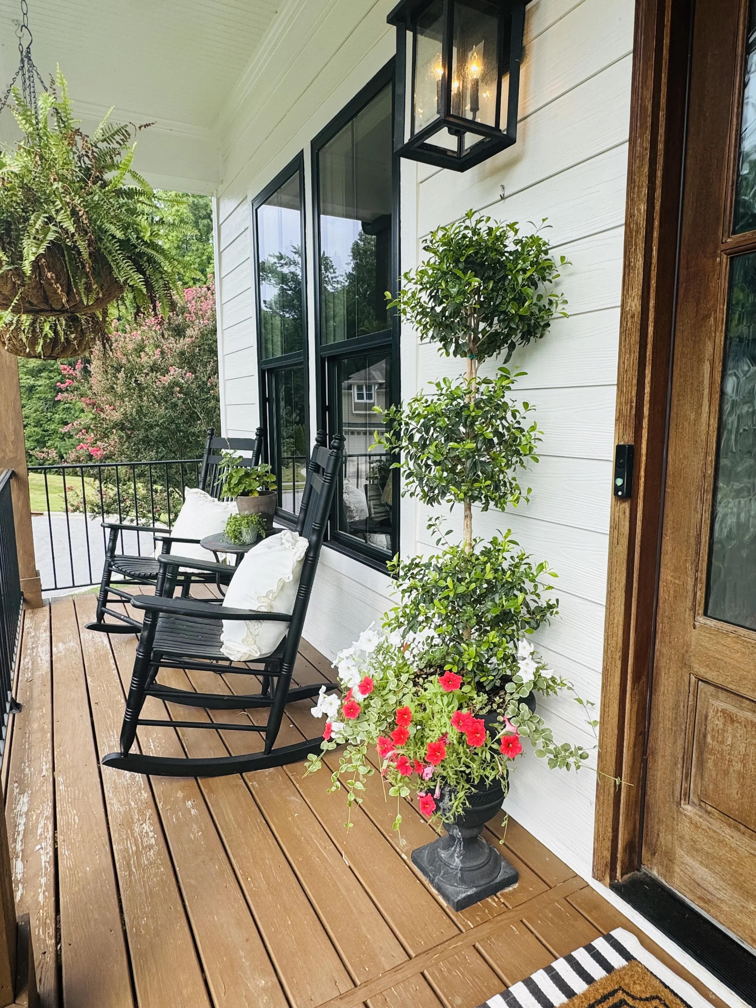 A porch with wooden flooring, black rocking chairs with white pillows, a large potted plant with pink and white flowers, a wall-mounted lantern light, white siding, and a wooden door.
