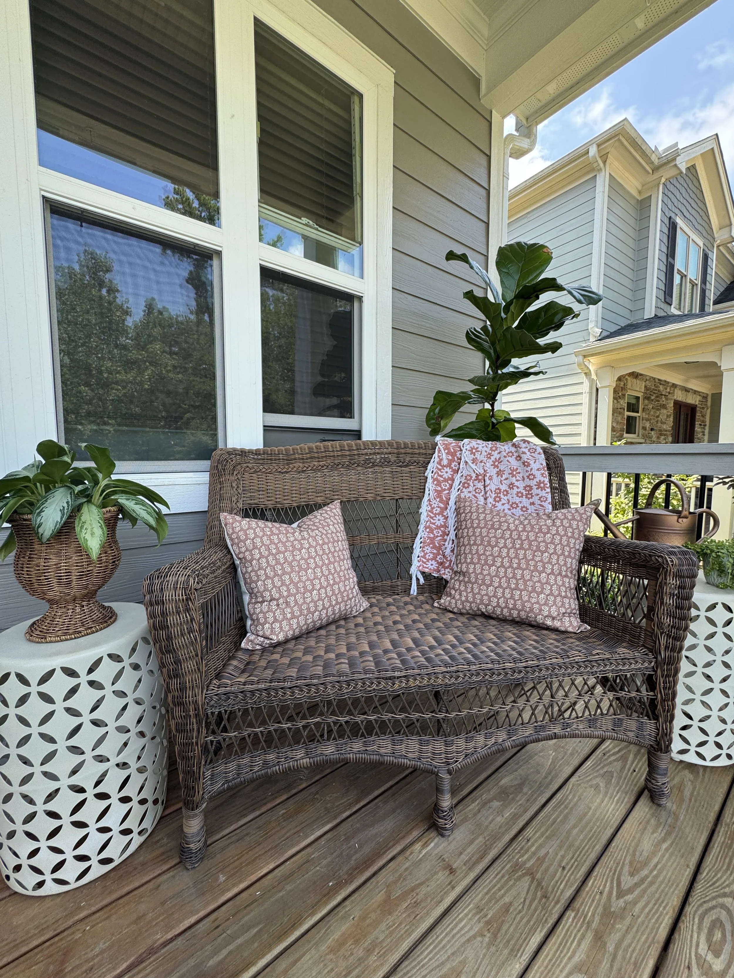 A wicker outdoor sofa with patterned pink cushions and pillows on a wooden deck. There is a white side table with a plant in a woven basket and a watering can. The house in the background has beige siding and large windows reflected with trees and blue sky.